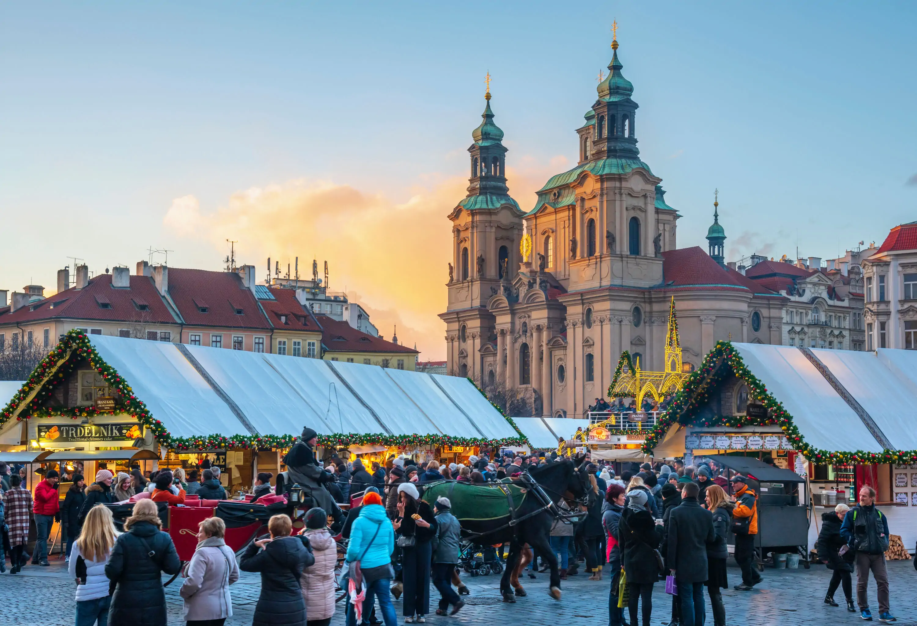 A crowd of people in the Christmas market with stalls decorated with Christmas ornaments, a horse with a red carriage, and the Baroque Church of Saint Nicholas in the background.