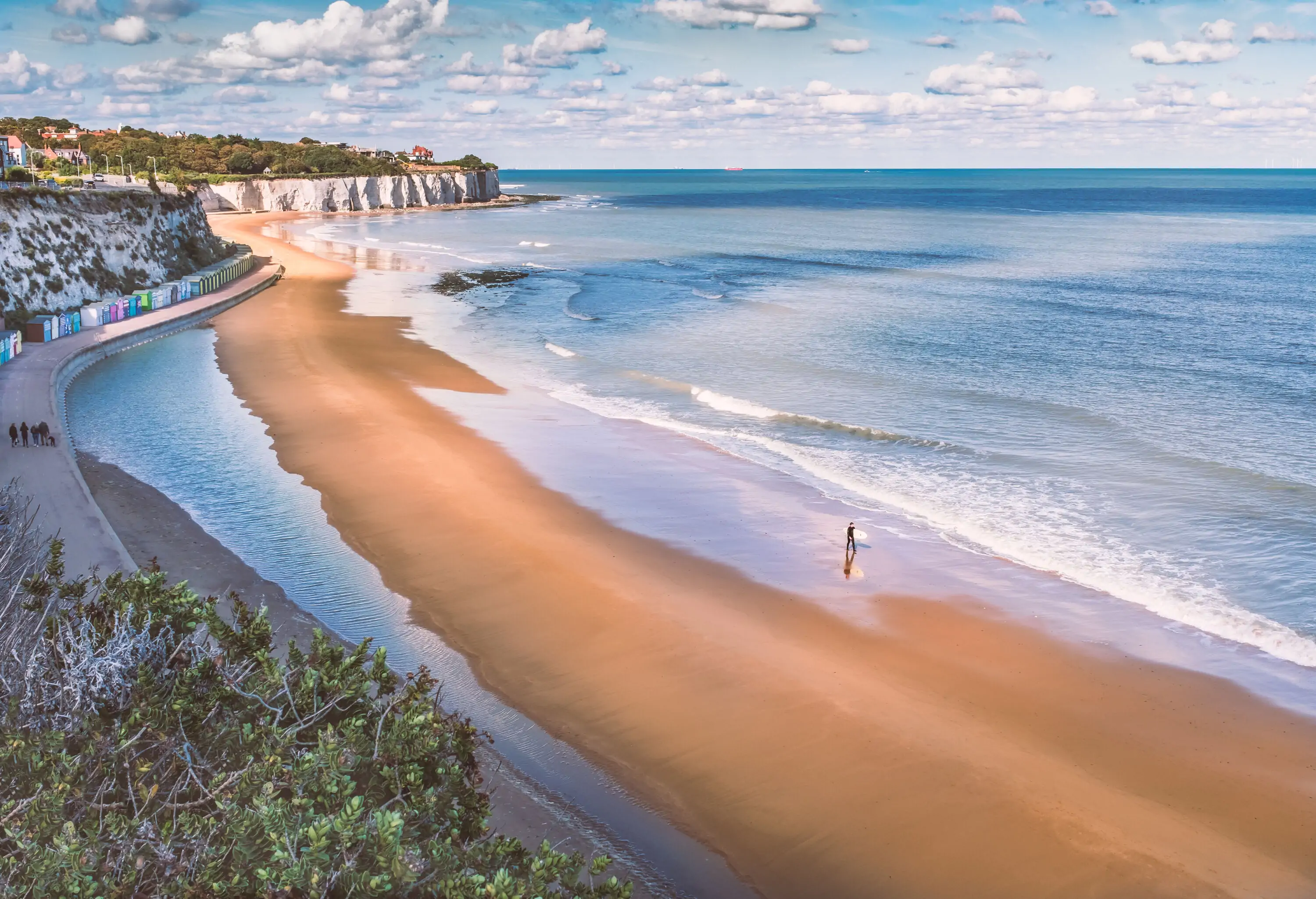 A lone surfer walks on a beach while holding his surfboard, with views of a distant white cliffs and a big ocean on the horizon.