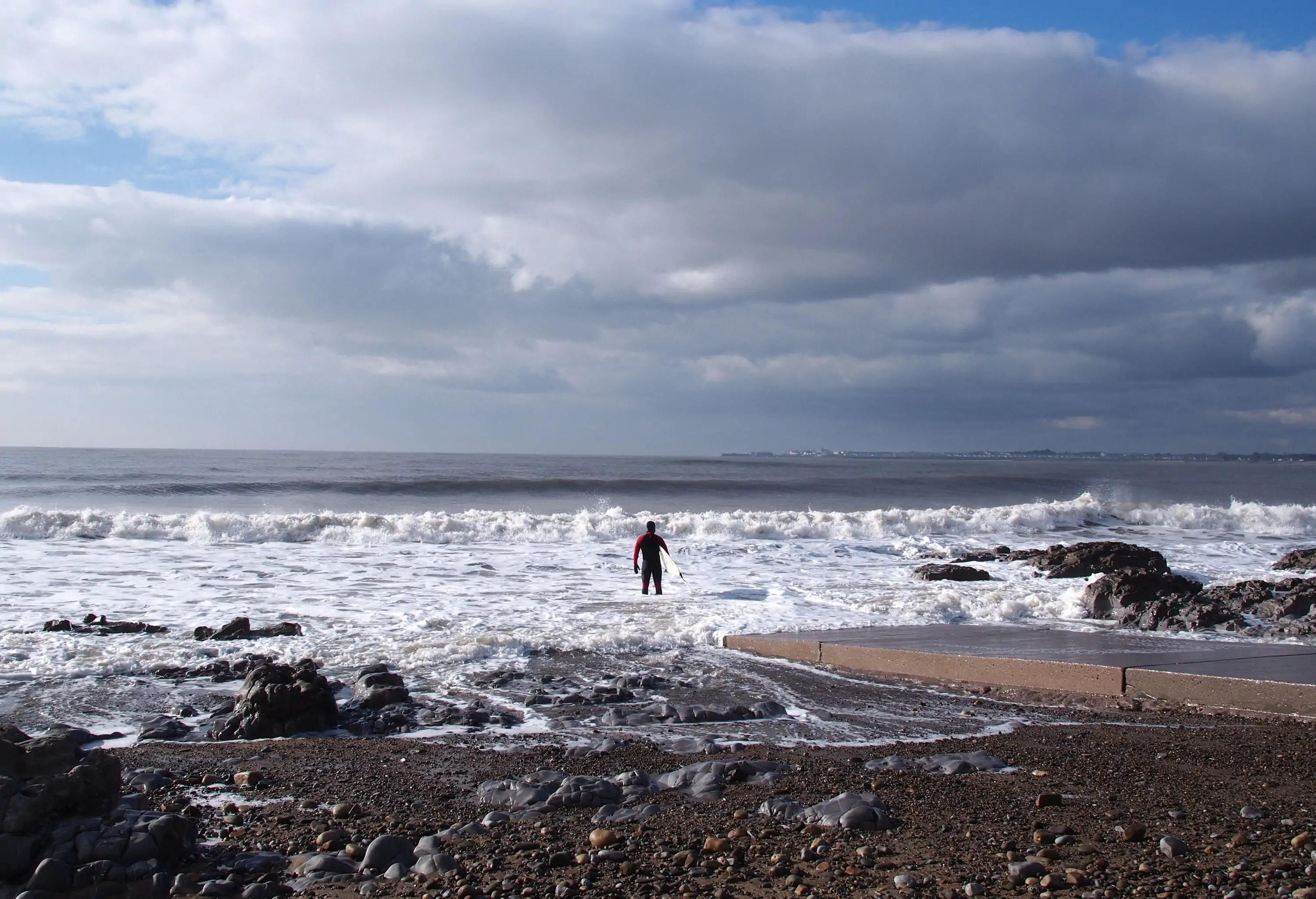 A male surfer standing on the shoreface as waves wash up on it.
