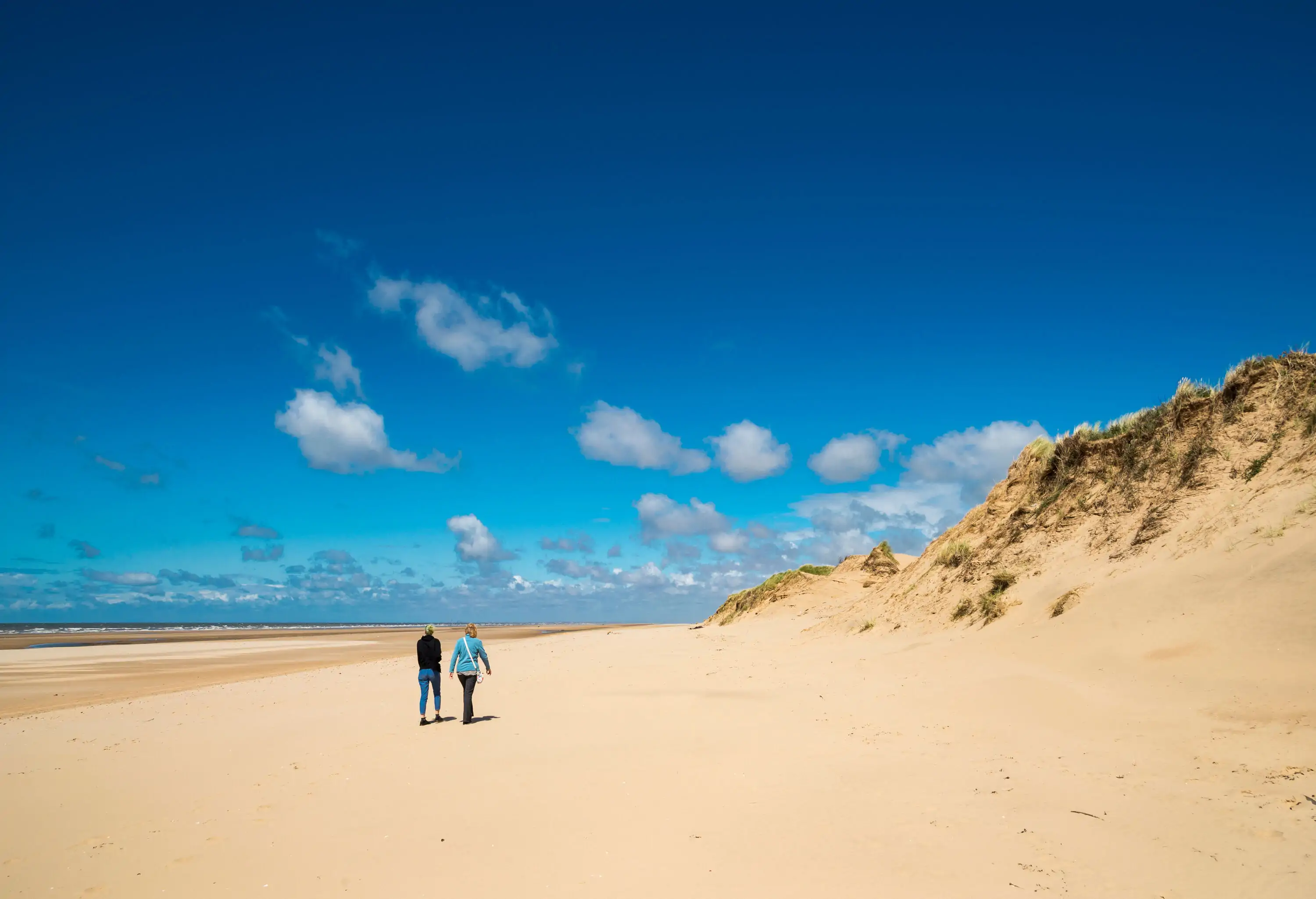 Two people enjoying a stroll along the sandy beach on the coast of Northwest England beneath a deep blue summer sky.