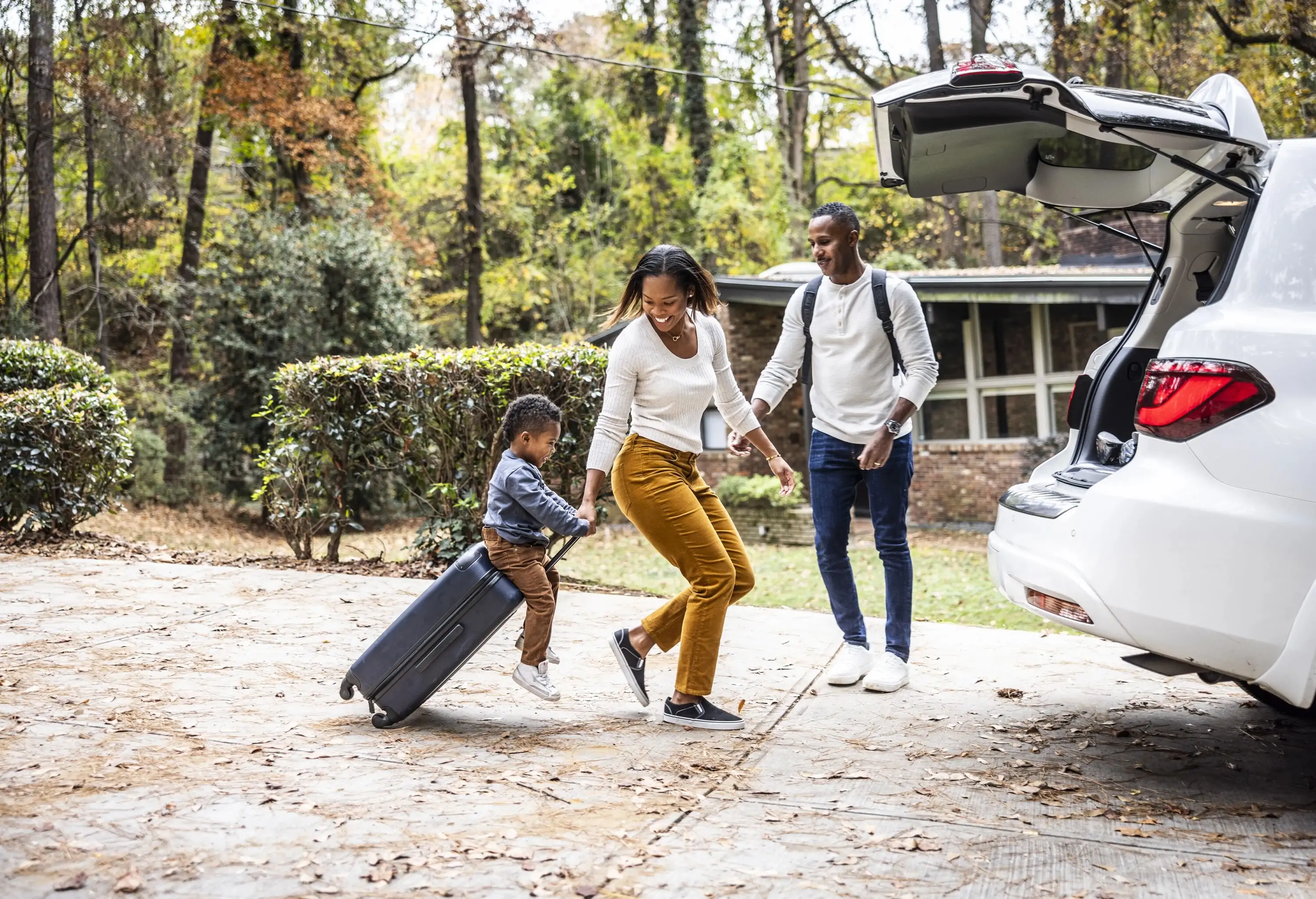 Mother pulling a young boy sitting on a suitcase while loading luggage into a car, preparing for a family vacation.