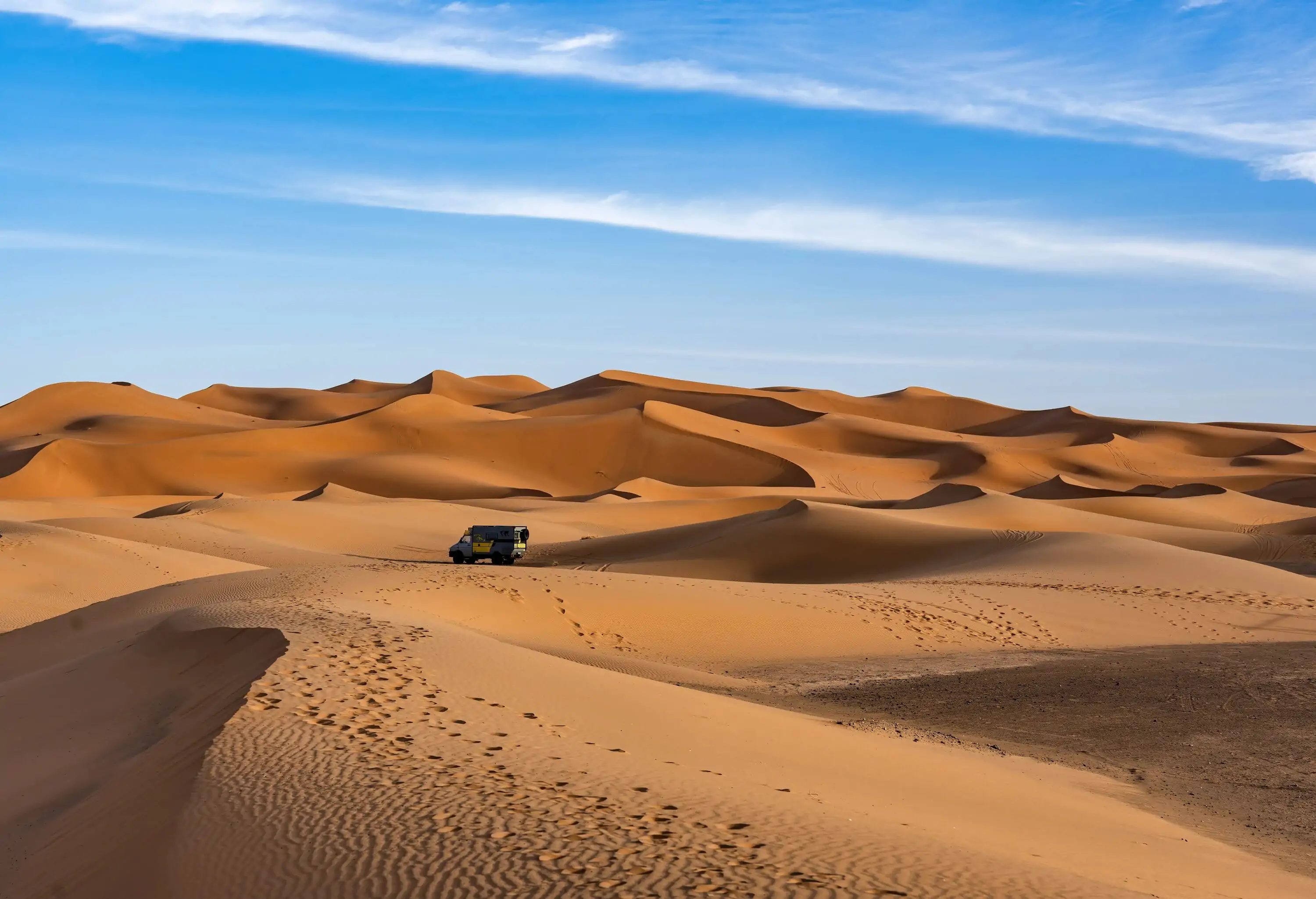 An off-road vehicle driving across the vast, golden dunes of the Sahara Desert under a wide blue sky, capturing the essence of adventure travel.