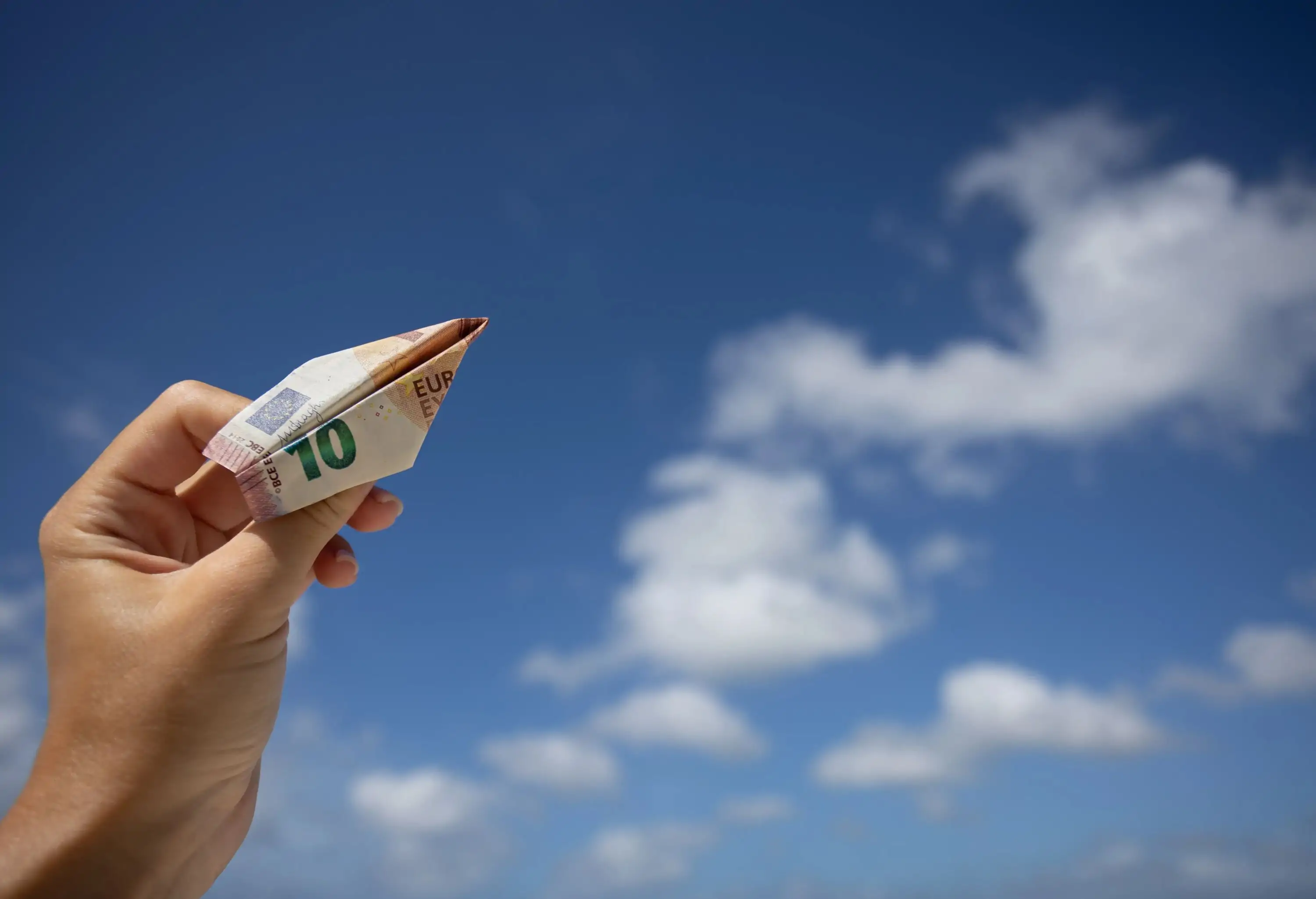 Hand holding the plane made of 10 euros bill on a blue sky with clouds background