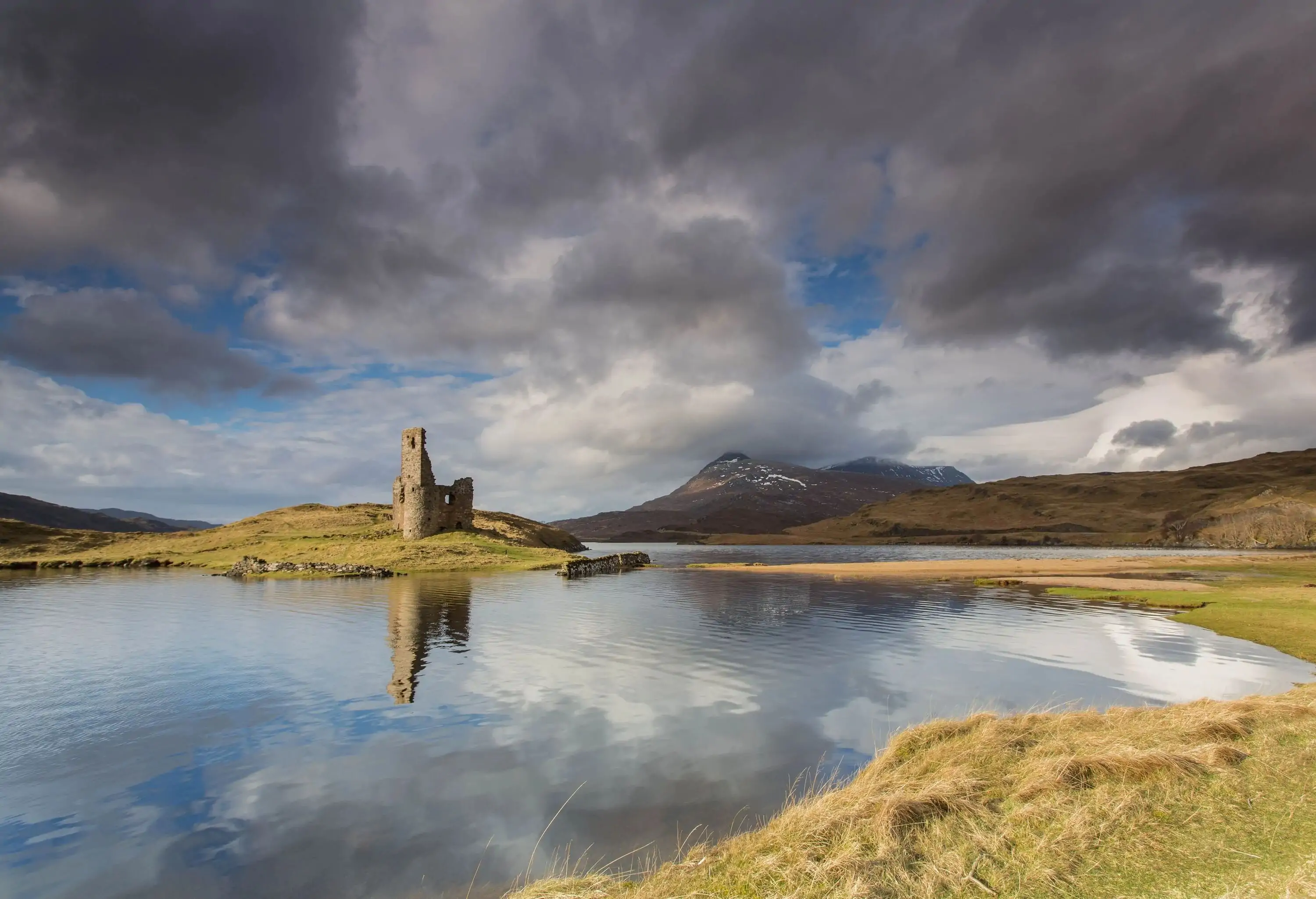 In the midst of a serene and verdant landscape, the ruined Ardvreck Castle stands as a haunting reminder of bygone days, its weathered walls and turrets casting their reflection upon the still waters below.