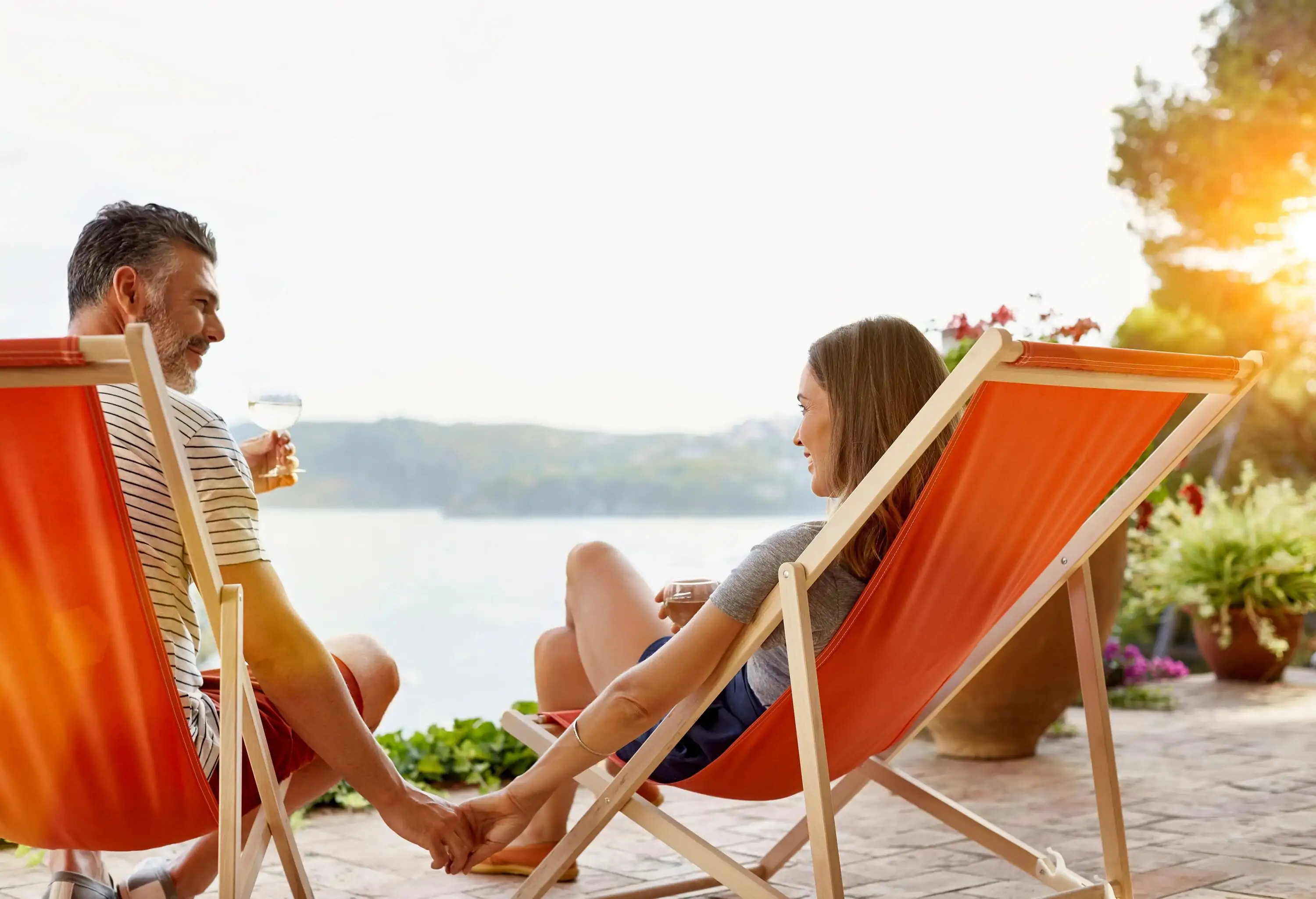 A couple relaxing on deck chairs on a patio with a magnificent view of the outdoors, drinking wine and holding hands.