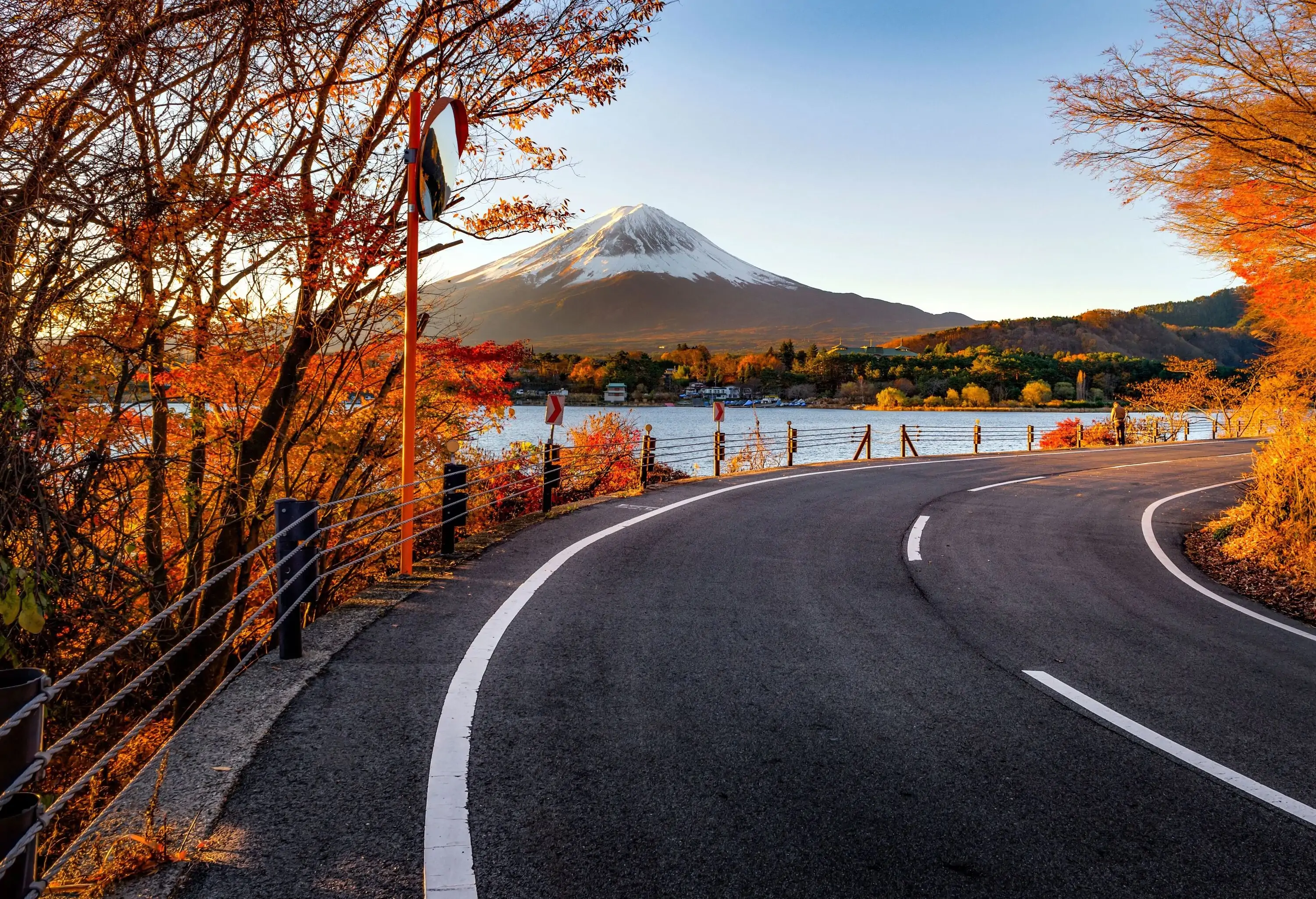 An empty road overlooking Mount Fuji with snow capped peak, surrounded by warm coloured autumn foliage on a crisp sunny fall day