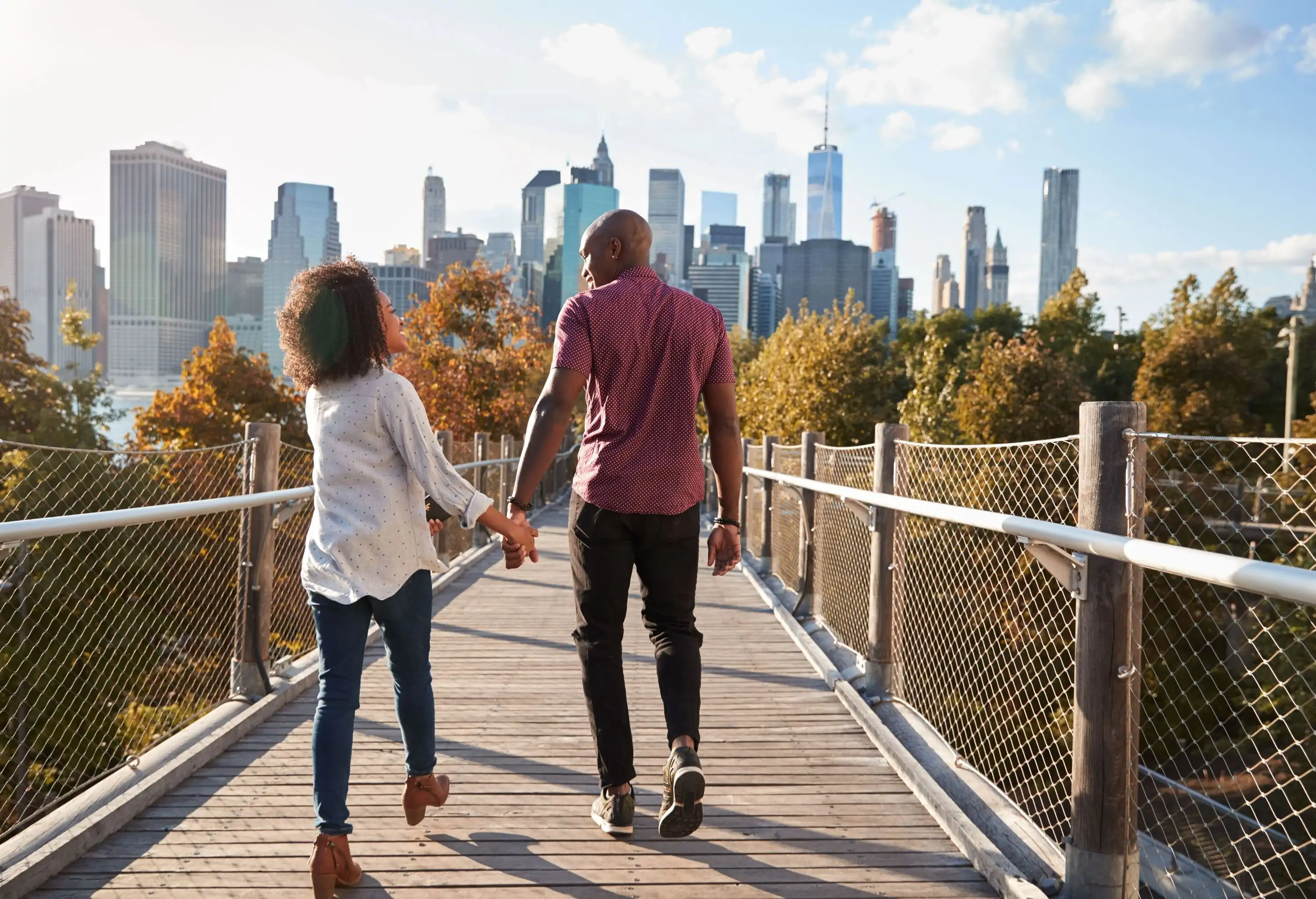 A couple holding hands as they walk through a bridge, with lofty skyscrapers in the background.