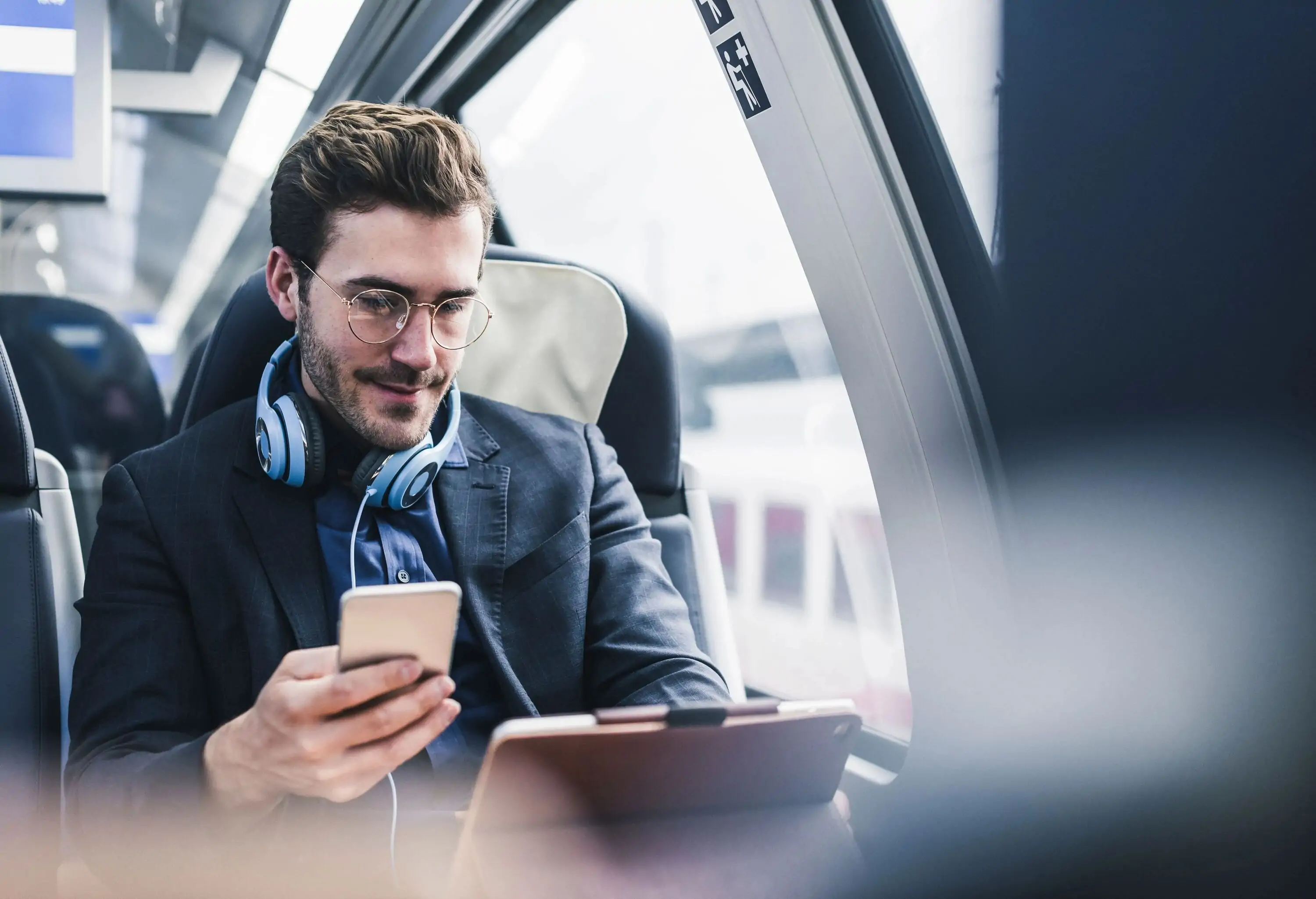 A businessman wearing blue headphones on his neck is working on an iPad while holding a phone inside a train.