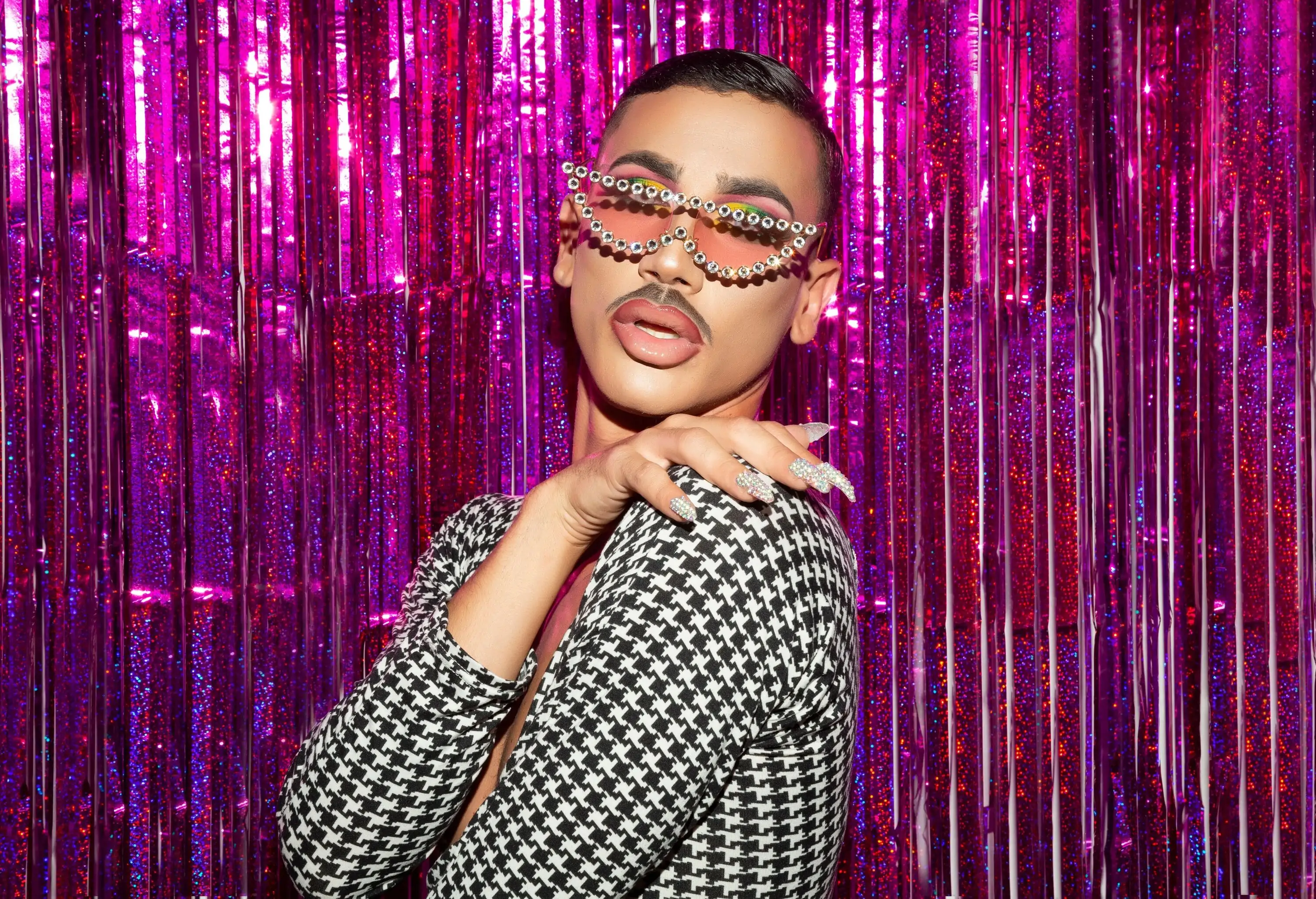 A drag queen in black and white chequered suit and decorated sunglasses poses against a shiny magenta background.