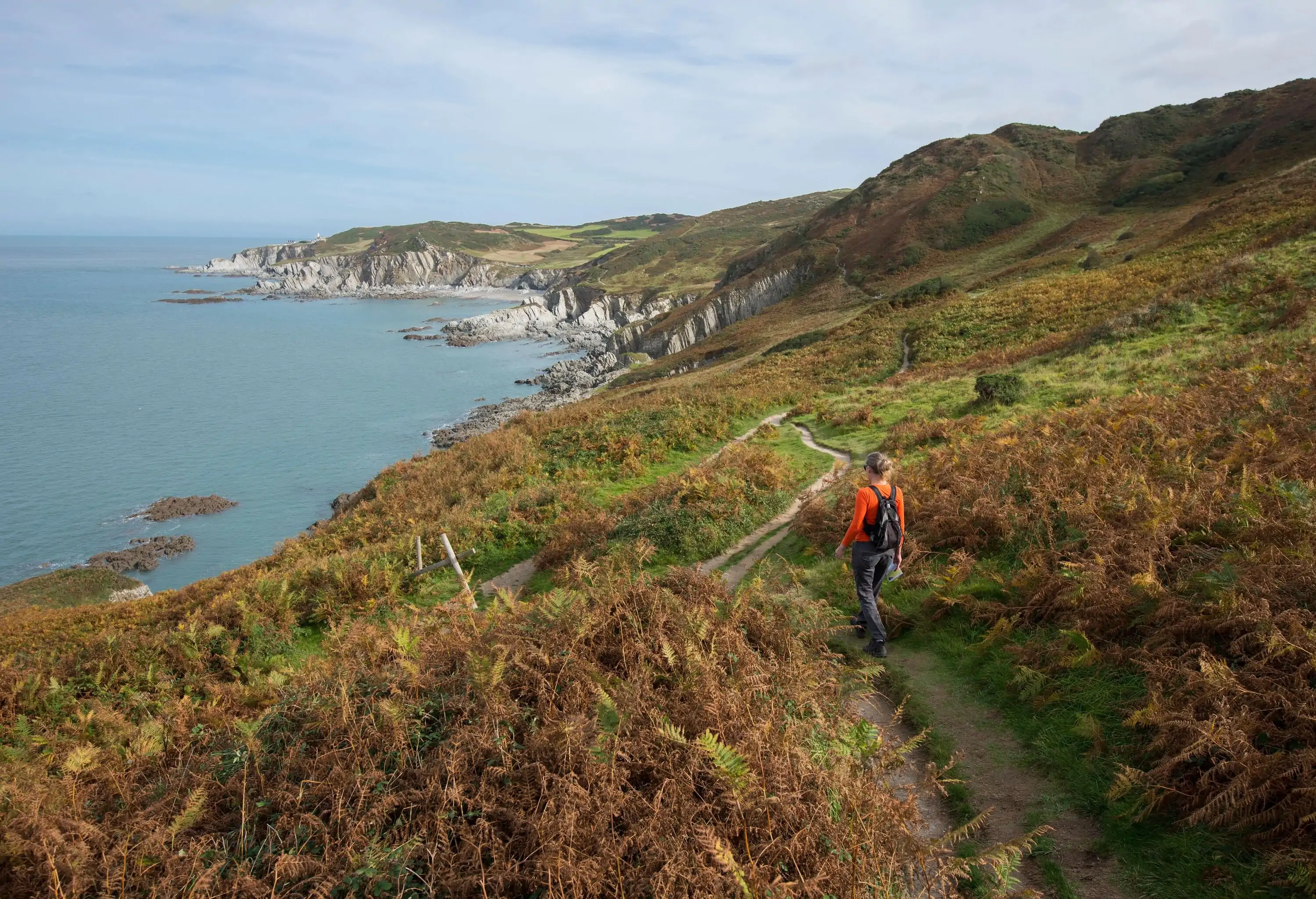 A woman in an orange top and small backpack walks along the South West Coast Path near Morte Point in North Devon in England UK