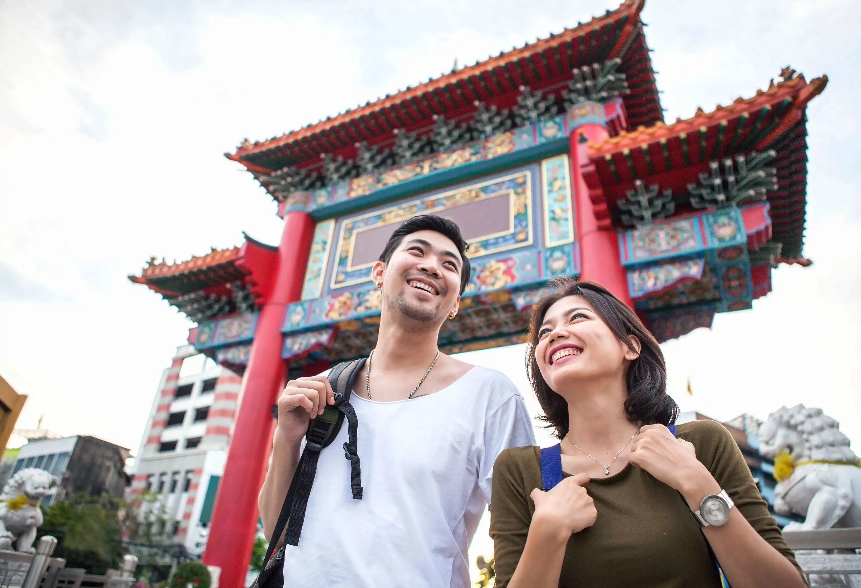 Couple in casual clothes smile while standing under a colourful traditional-style gateway.