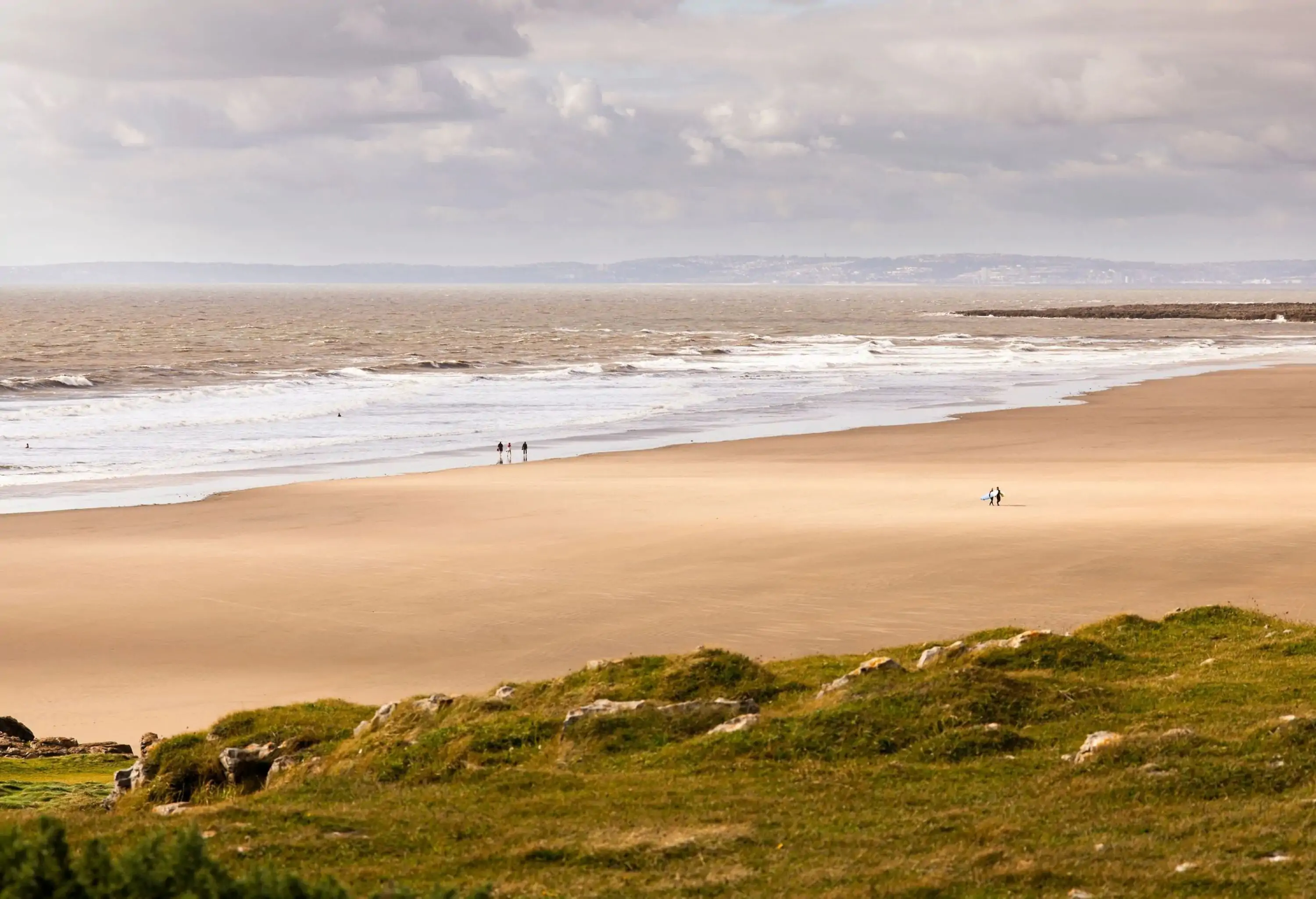 Wide sandy beach with people seen from afar from the top of a grassy mountain on an overcast windy day. Rest Bay, South Wales UK