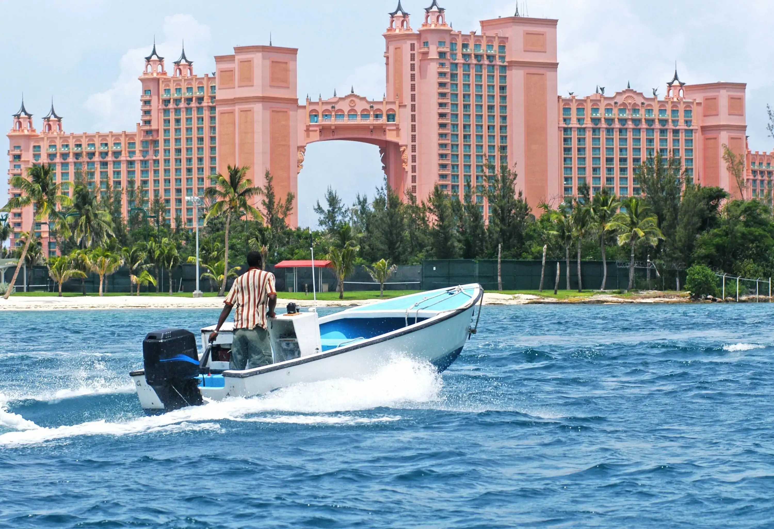 A man drives a speeding boat on a beach with views of a massive hotel with a bridge in middle connecting two buildings.