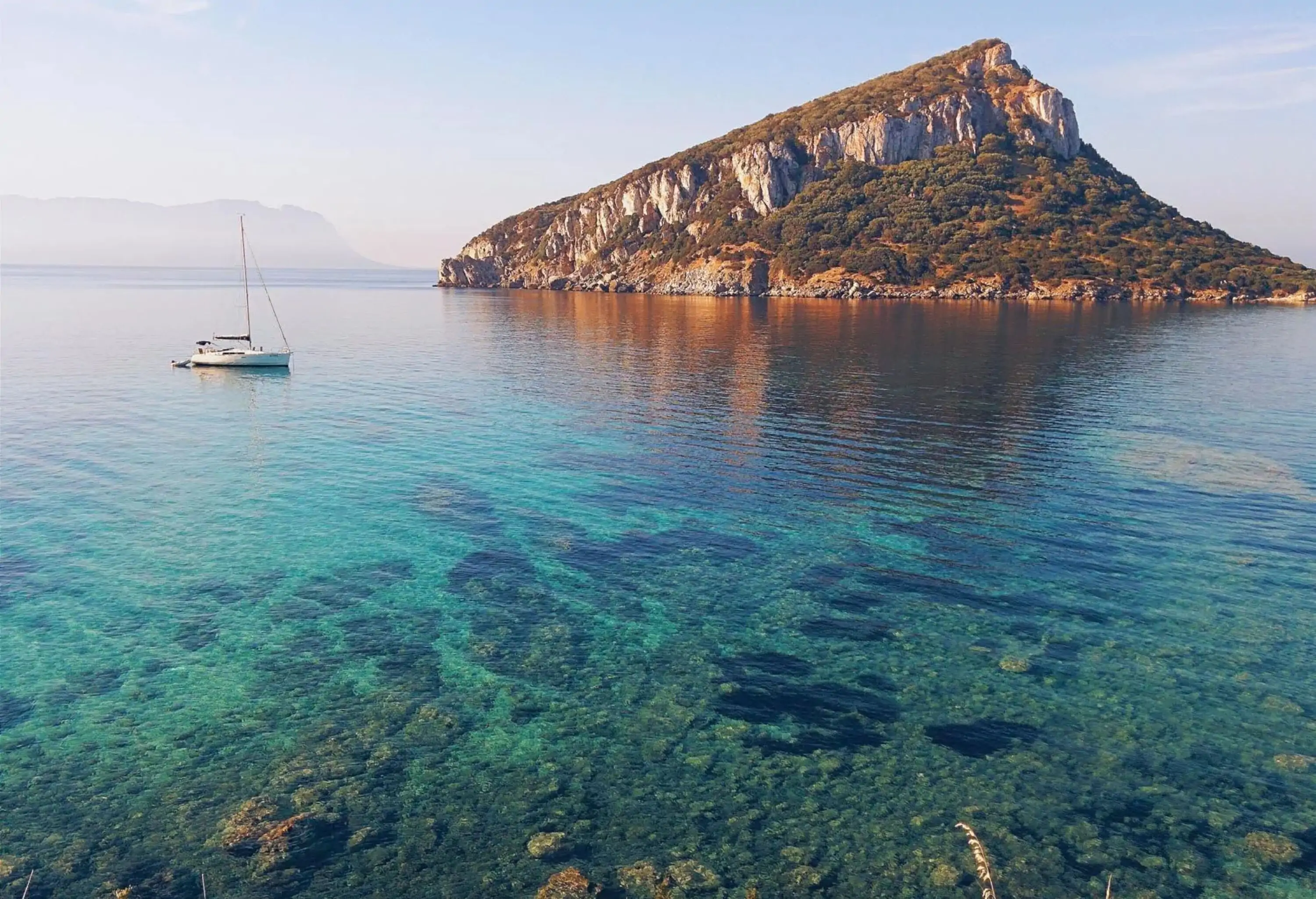 A small white boat moored near an imposing rock mountain surrounded by shallow crystalline water.