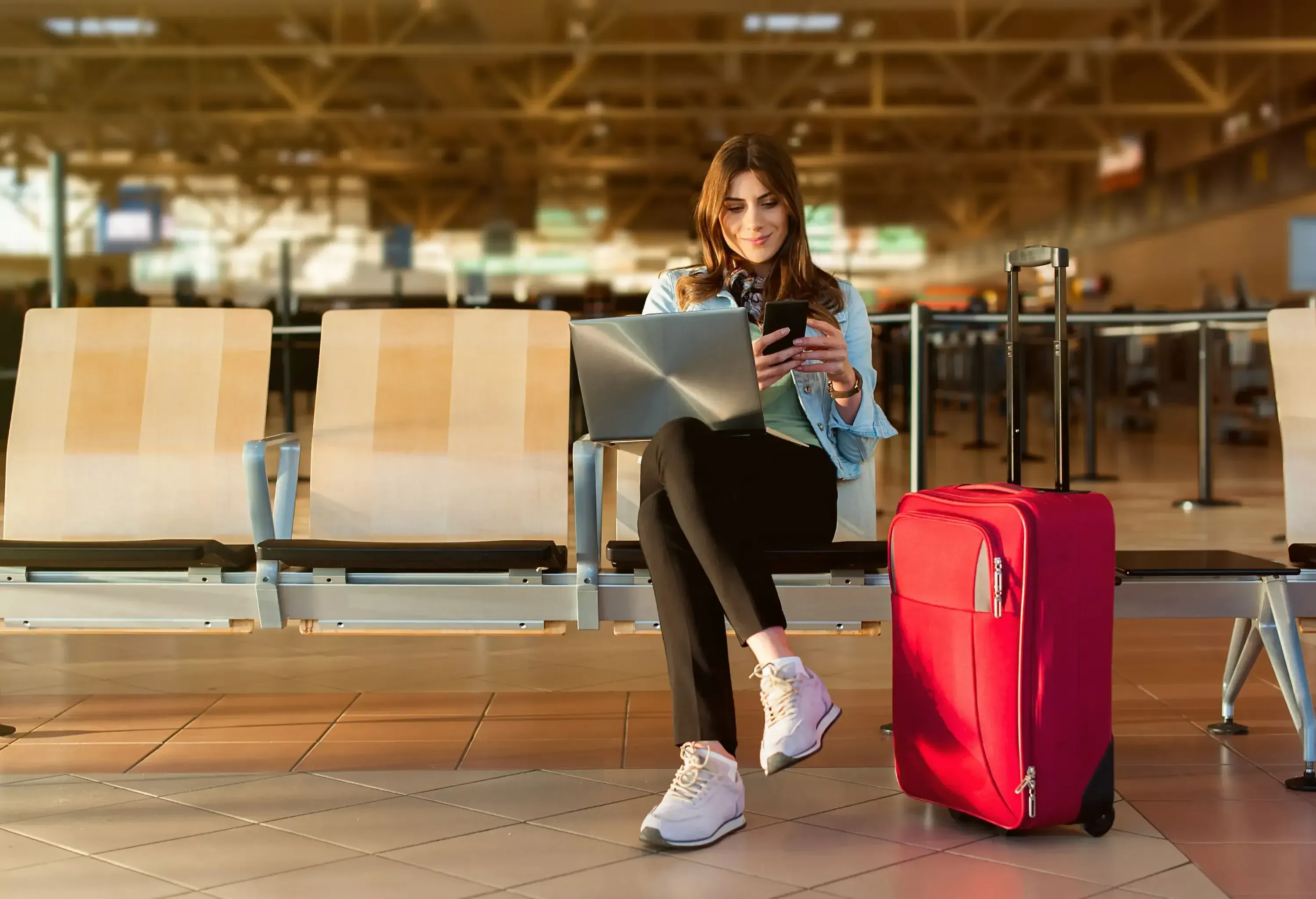 A woman using her laptop and phone while sitting next to her pink suitcase at an airport terminal.