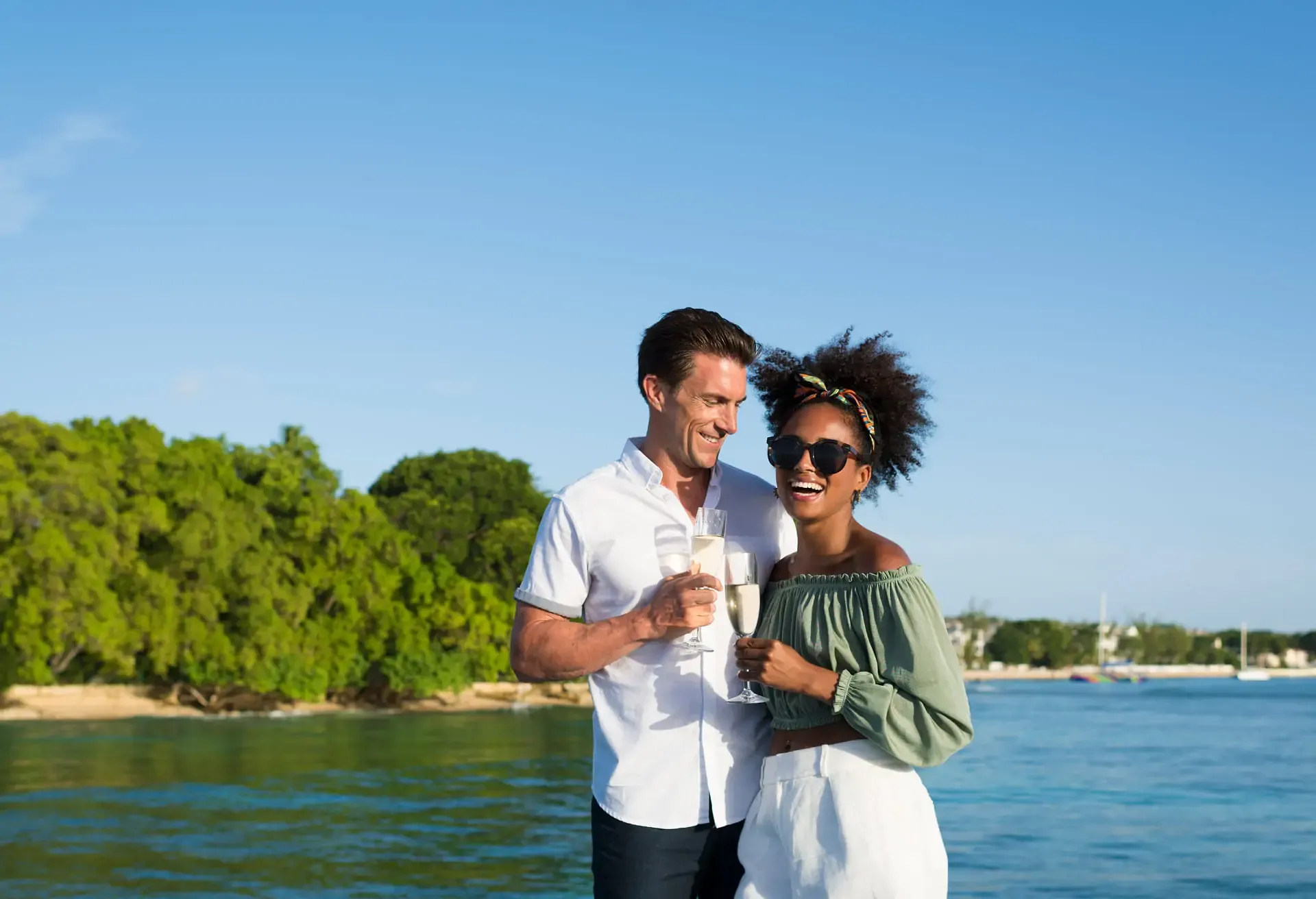 A happy couple holding champagne flutes by the water with a green island background.