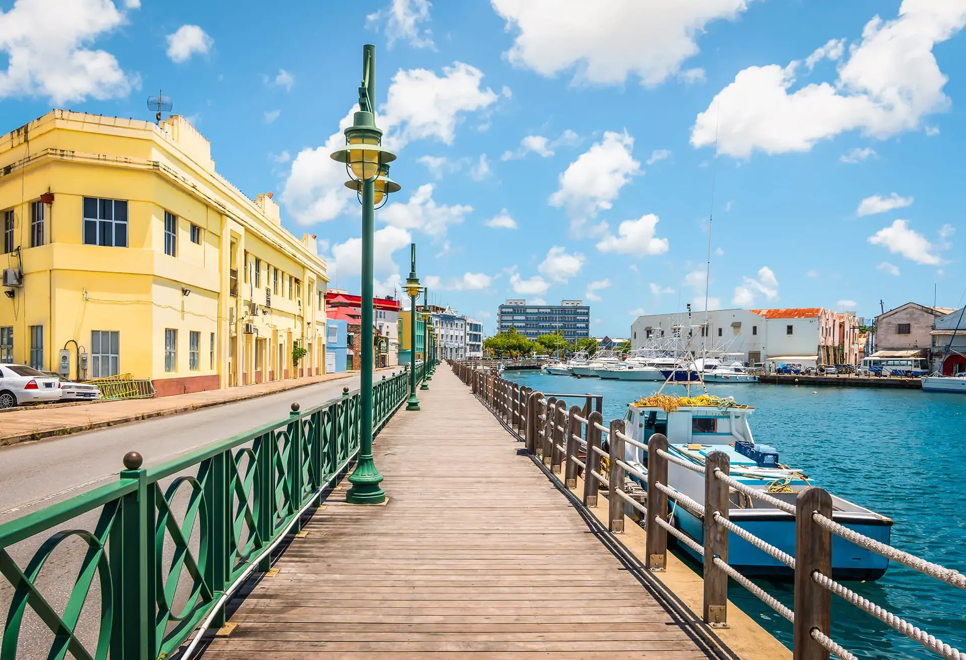 A sunny waterfront boardwalk with a yellow building, green railings, and boats on the water.