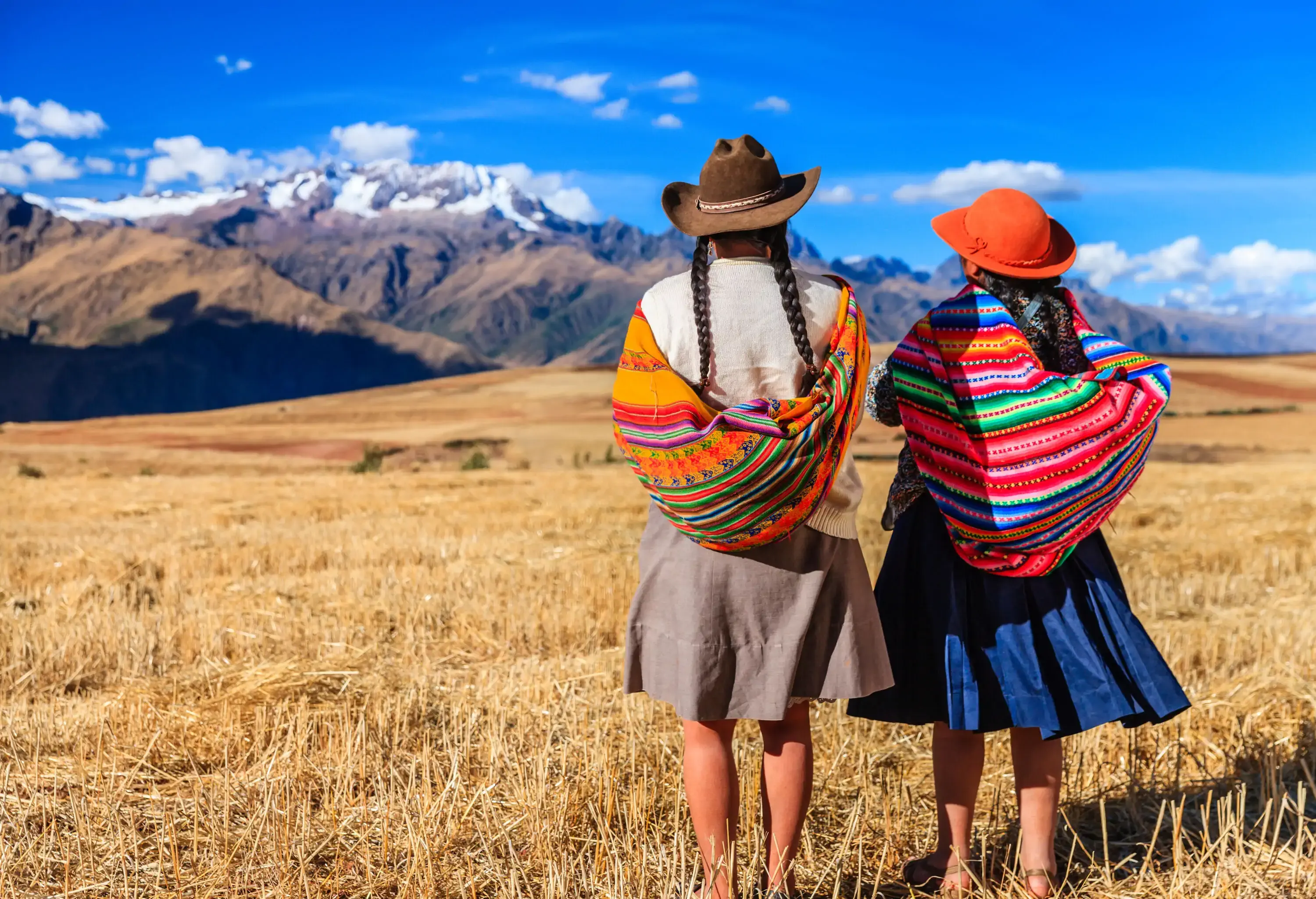 Two women in colourful shawls, hats, and skirts stand on a grass field as they look at snow-capped mountains.