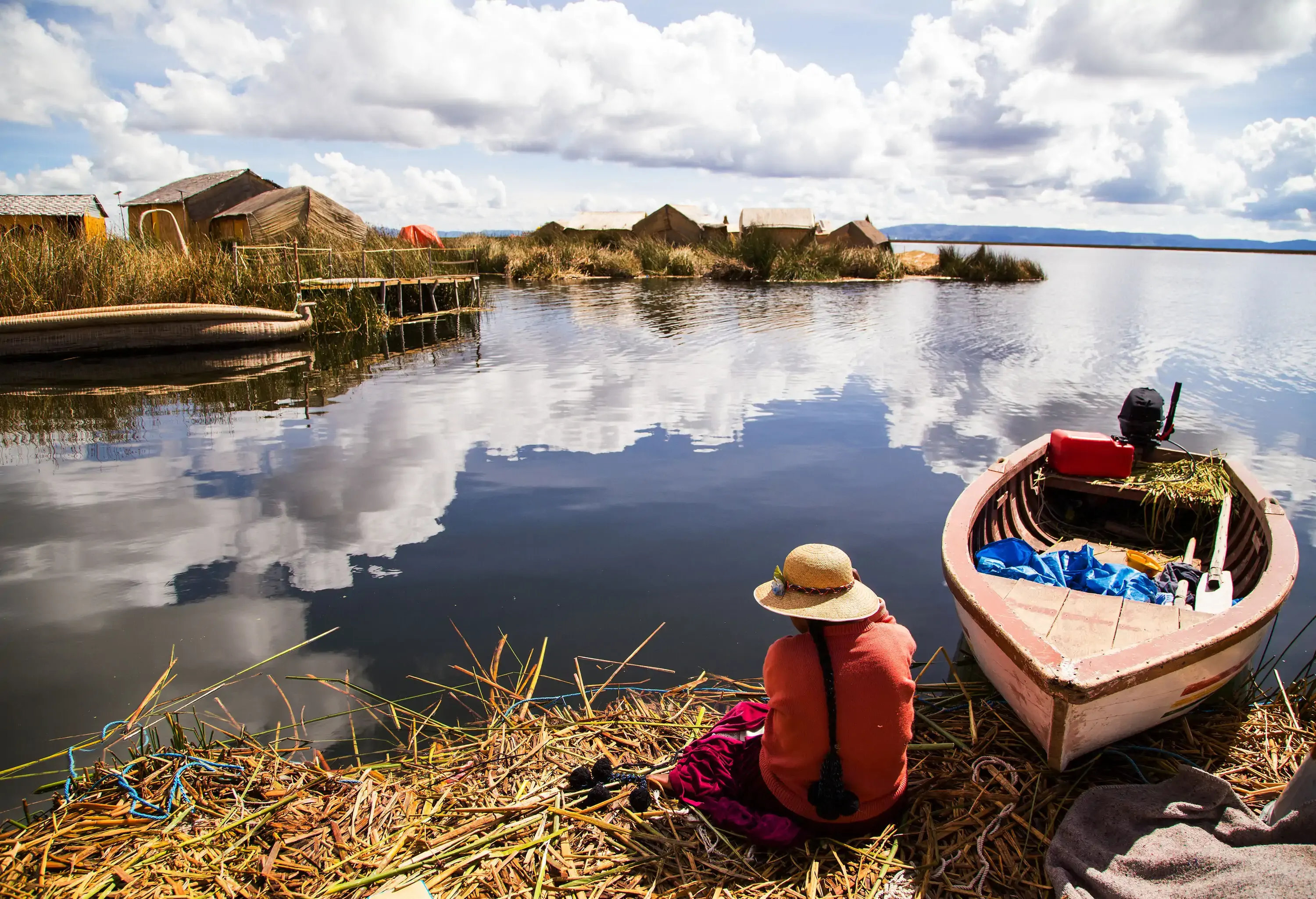 A person in a hat sits on a pile of floating twigs next to a boat on a nearby floating village.