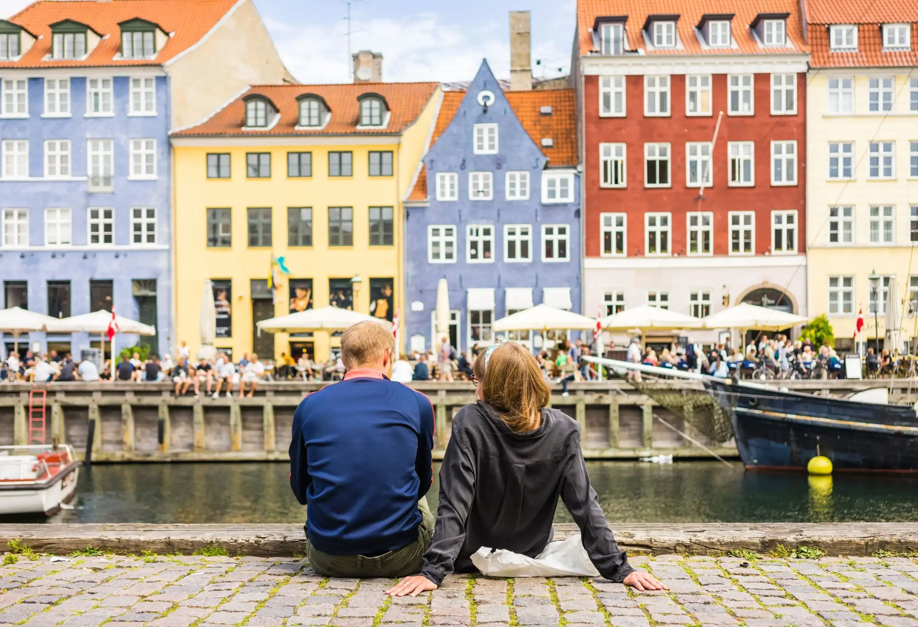 Two individuals sit on the promenade by the canal overlooking the pedestrians on the street lined with colourful and classic buildings.