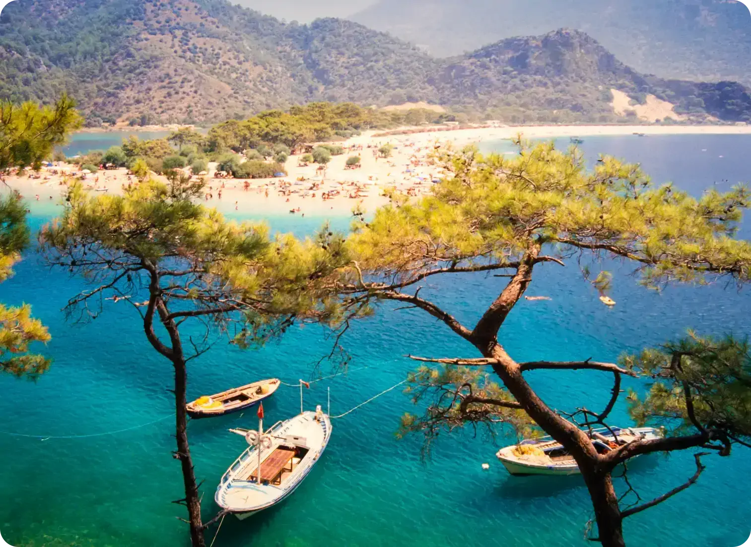 Scenic beach with turquoise waters, boats anchored near shore, pine trees, and mountains in the background.