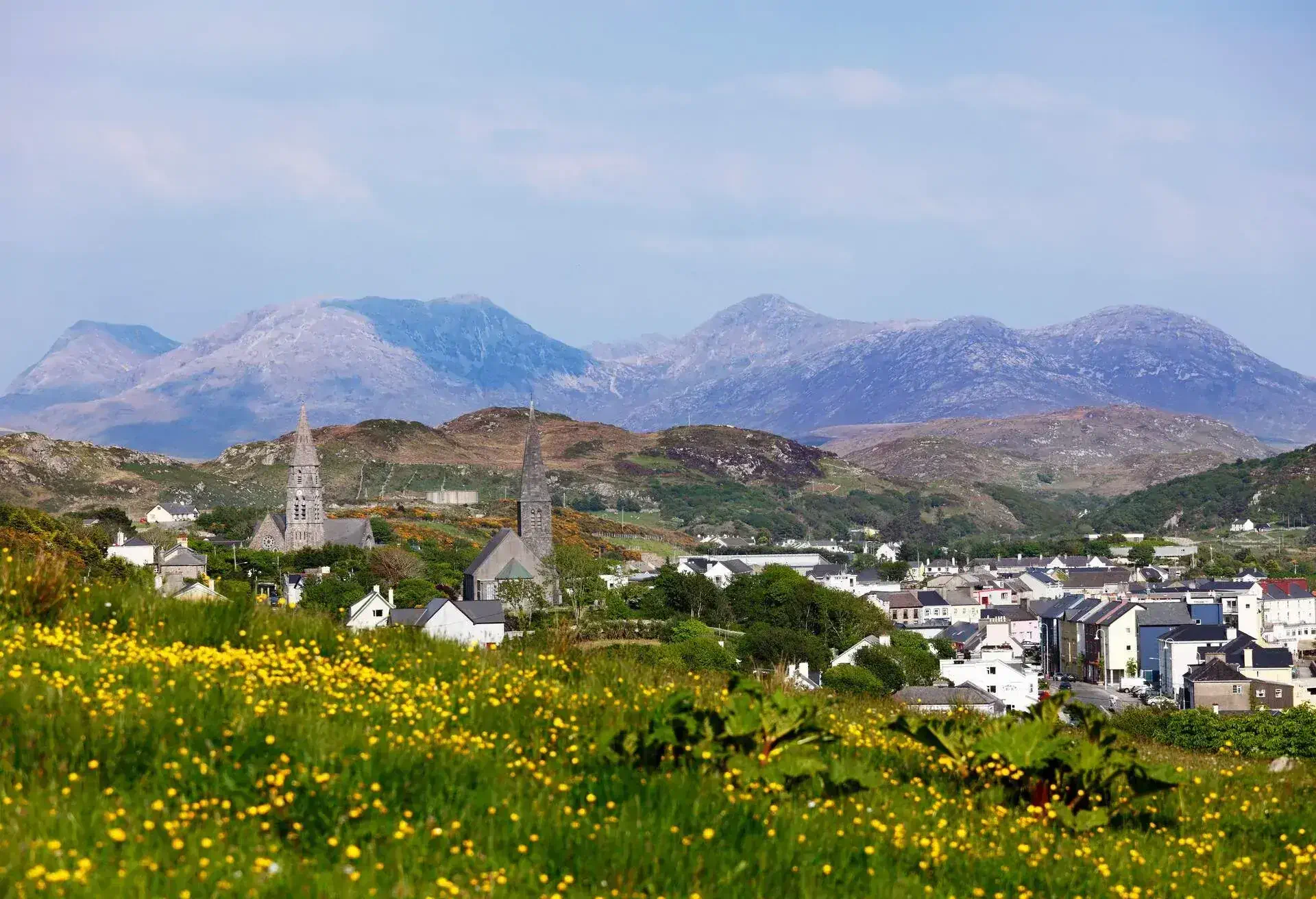 View of a small Irish town with stone church spires, surrounded by green hills and yellow wildflowers, with mountains in the background.
