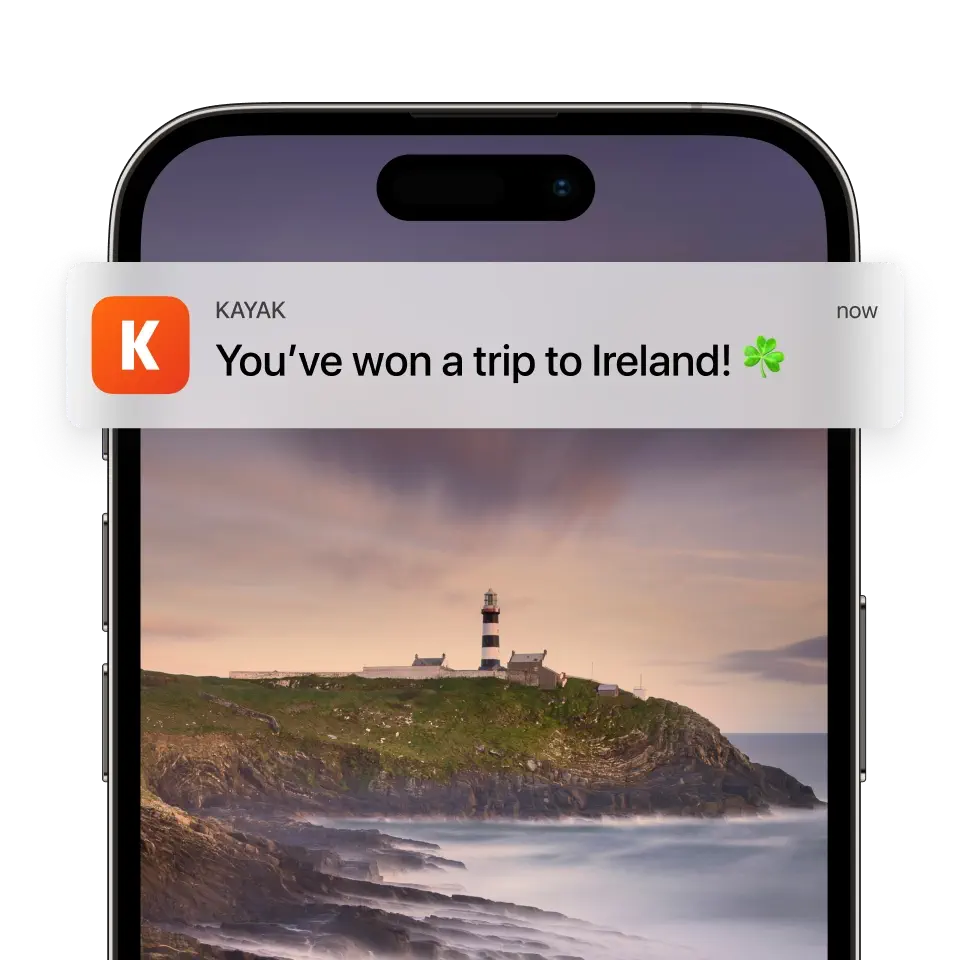Phone screen with a KAYAK notification: “You’ve won a trip to Ireland! ☘️” over a lighthouse on a rocky coast.
