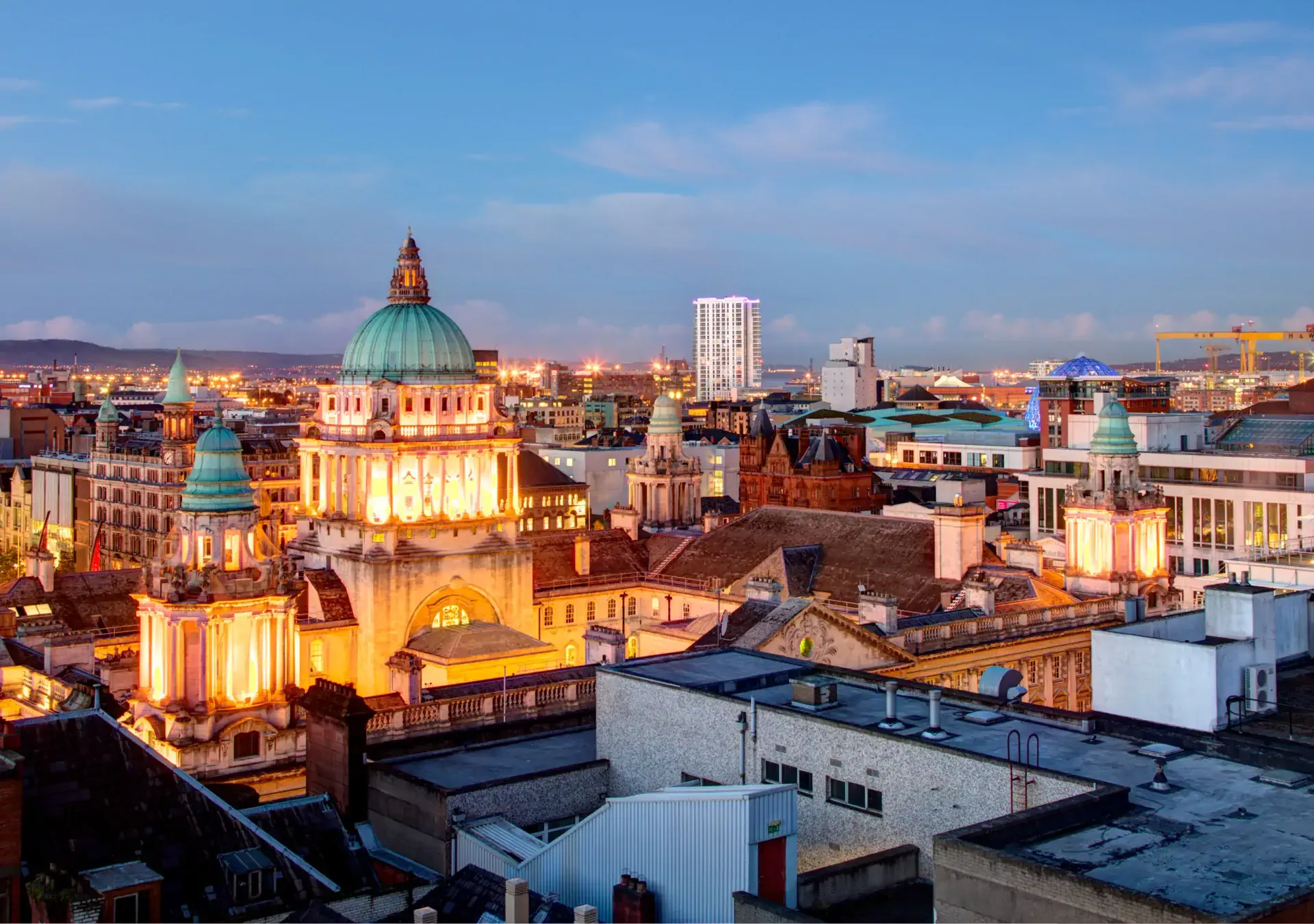 Evening view of Belfast City Hall with its green dome and illuminated facade, surrounded by city buildings and cranes in the distance.