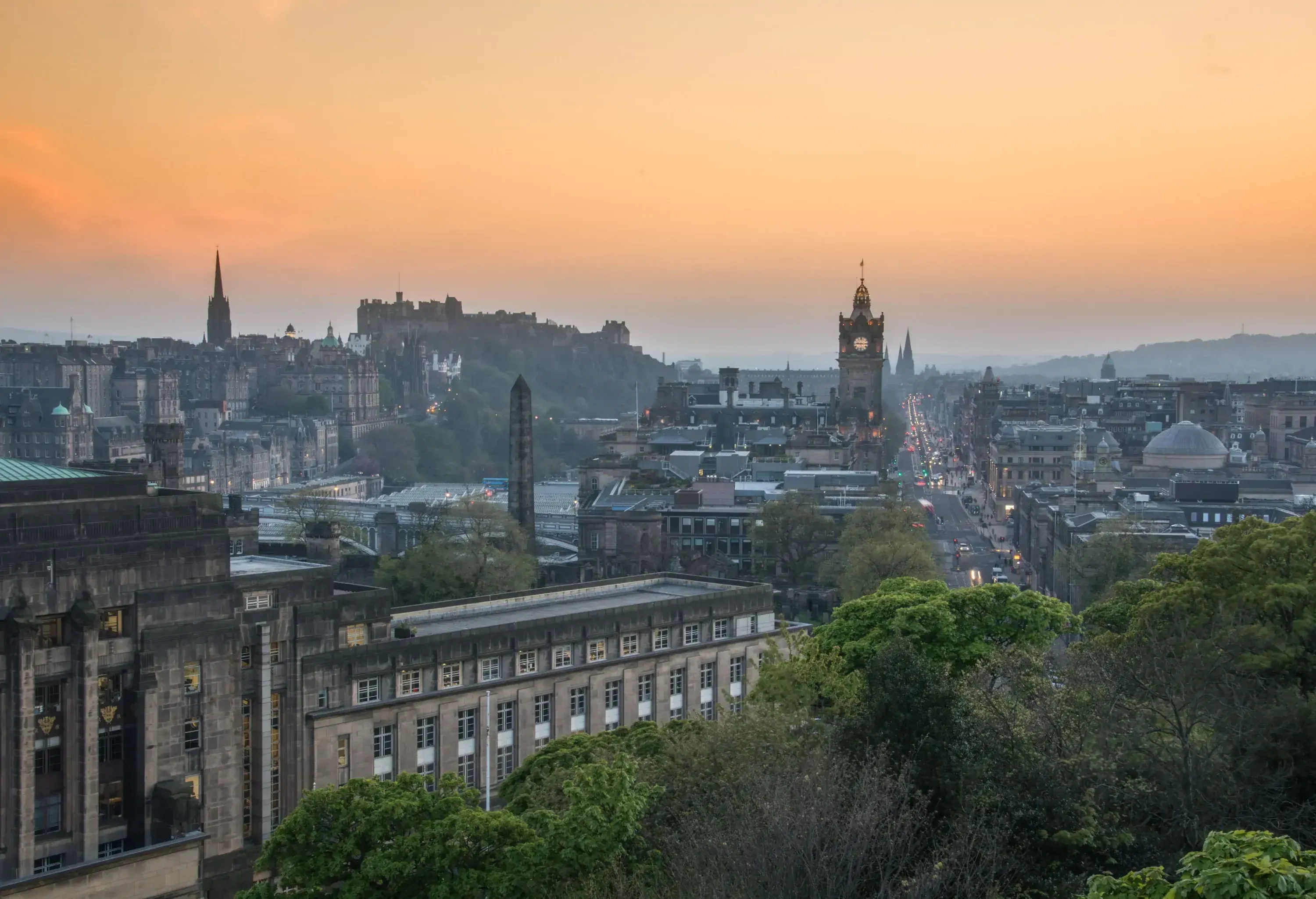 A clock tower rises above a cluster of old city buildings under an orange sky.