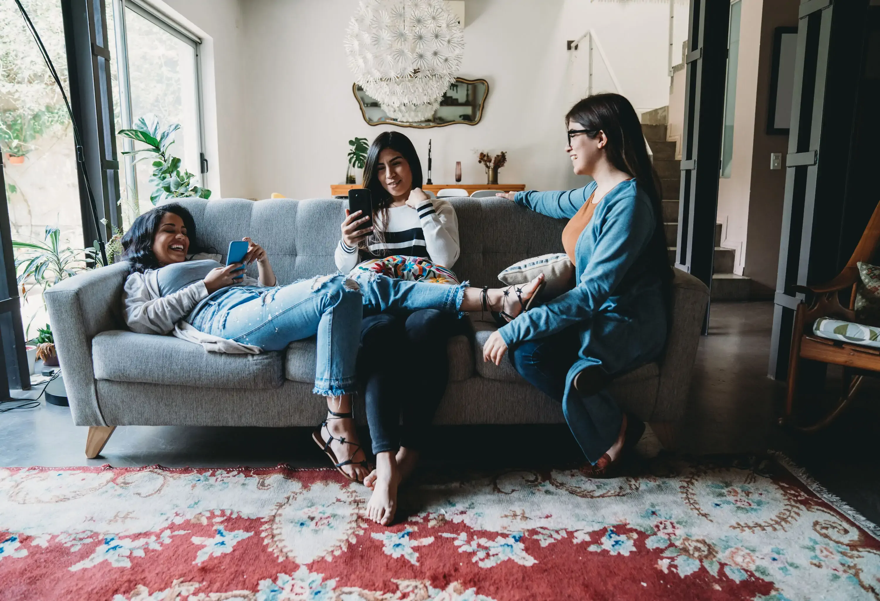Three female friends talk and hang out in the living room sofa while using their phones.