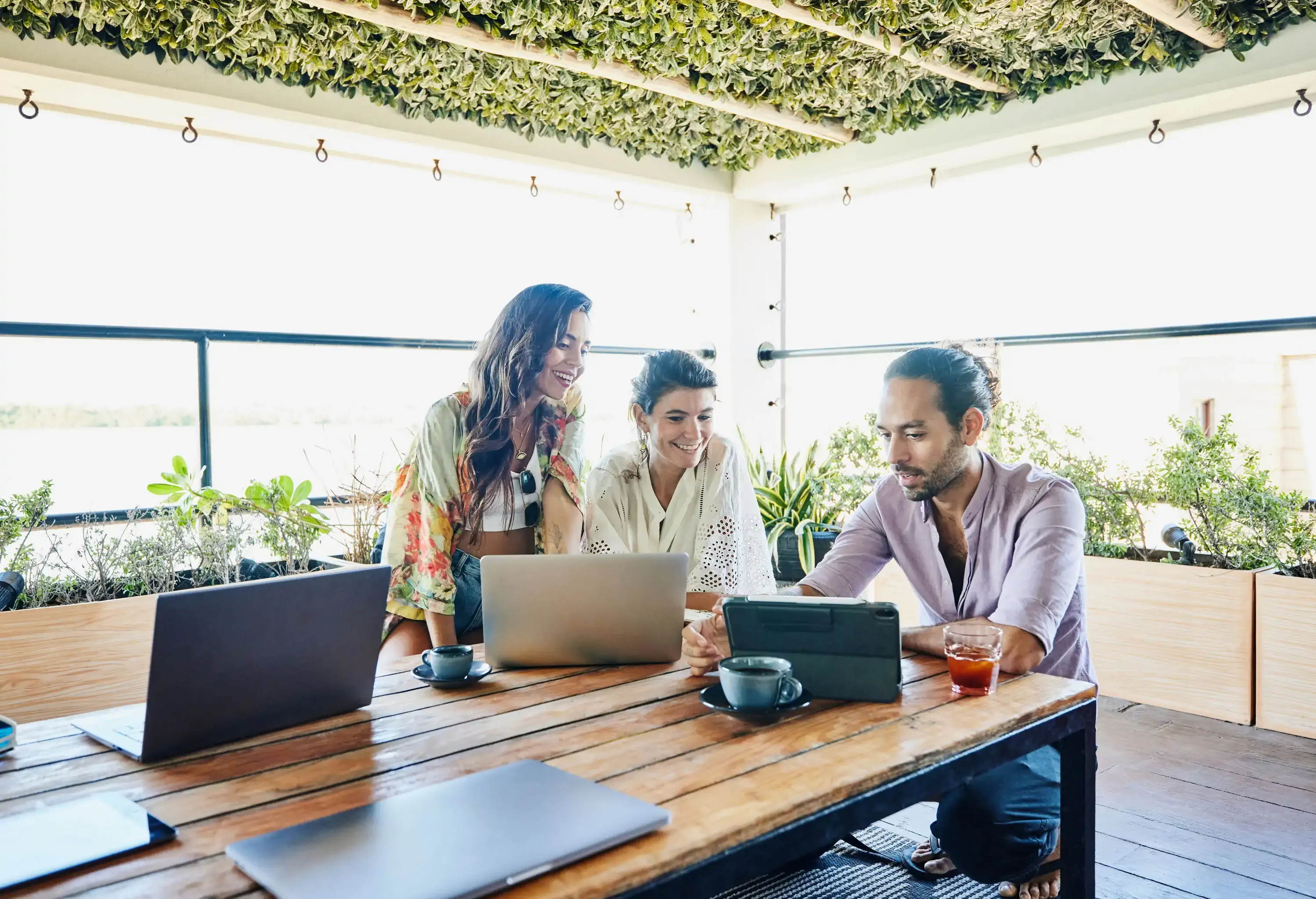 Medium wide shot of smiling colleagues discussing project on digital tablet while working remotely from deck of vacation rental