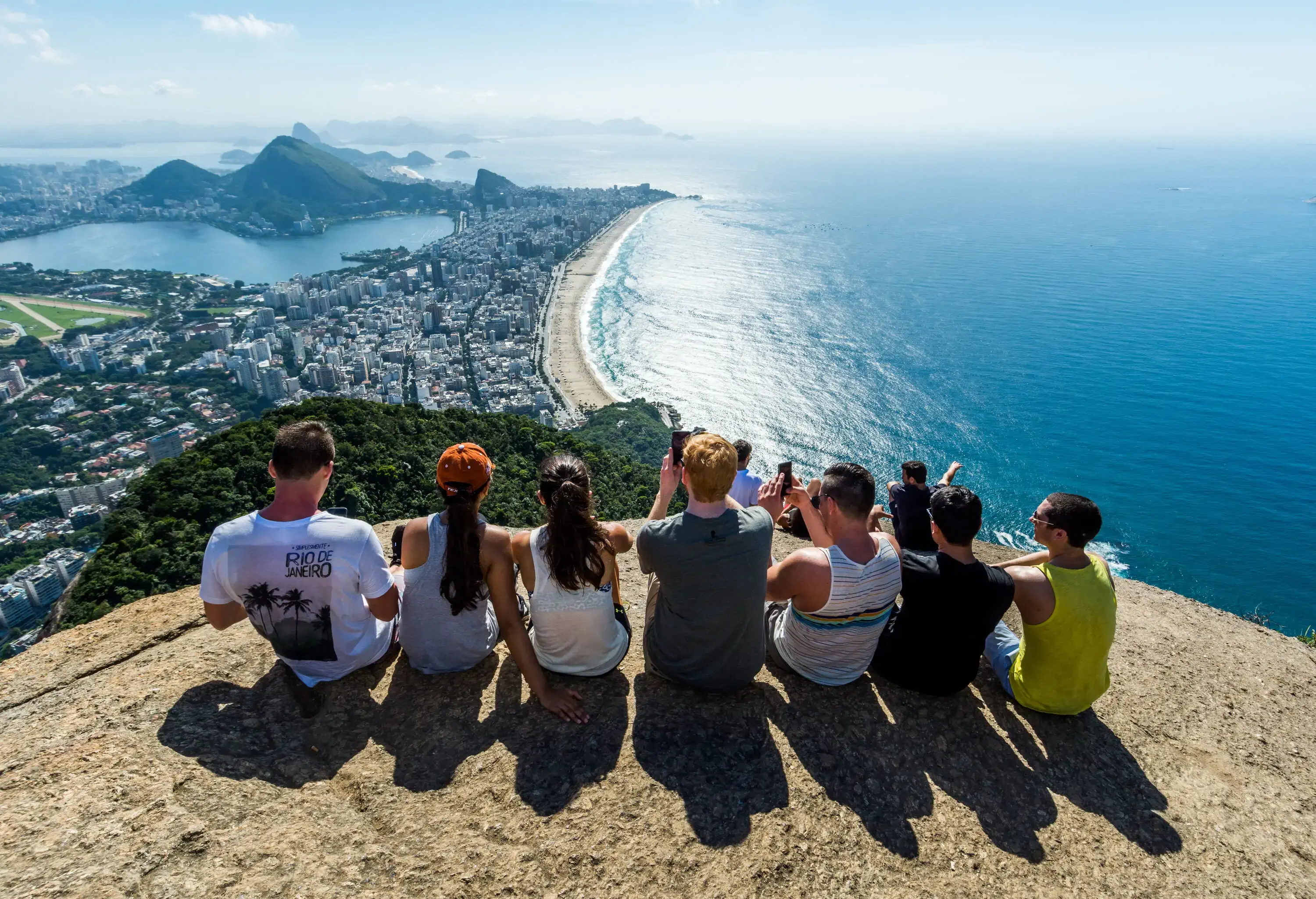 A group of friends sitting on a cliff and taking photos at the forest and urban landscape of a coastal metropolis below.