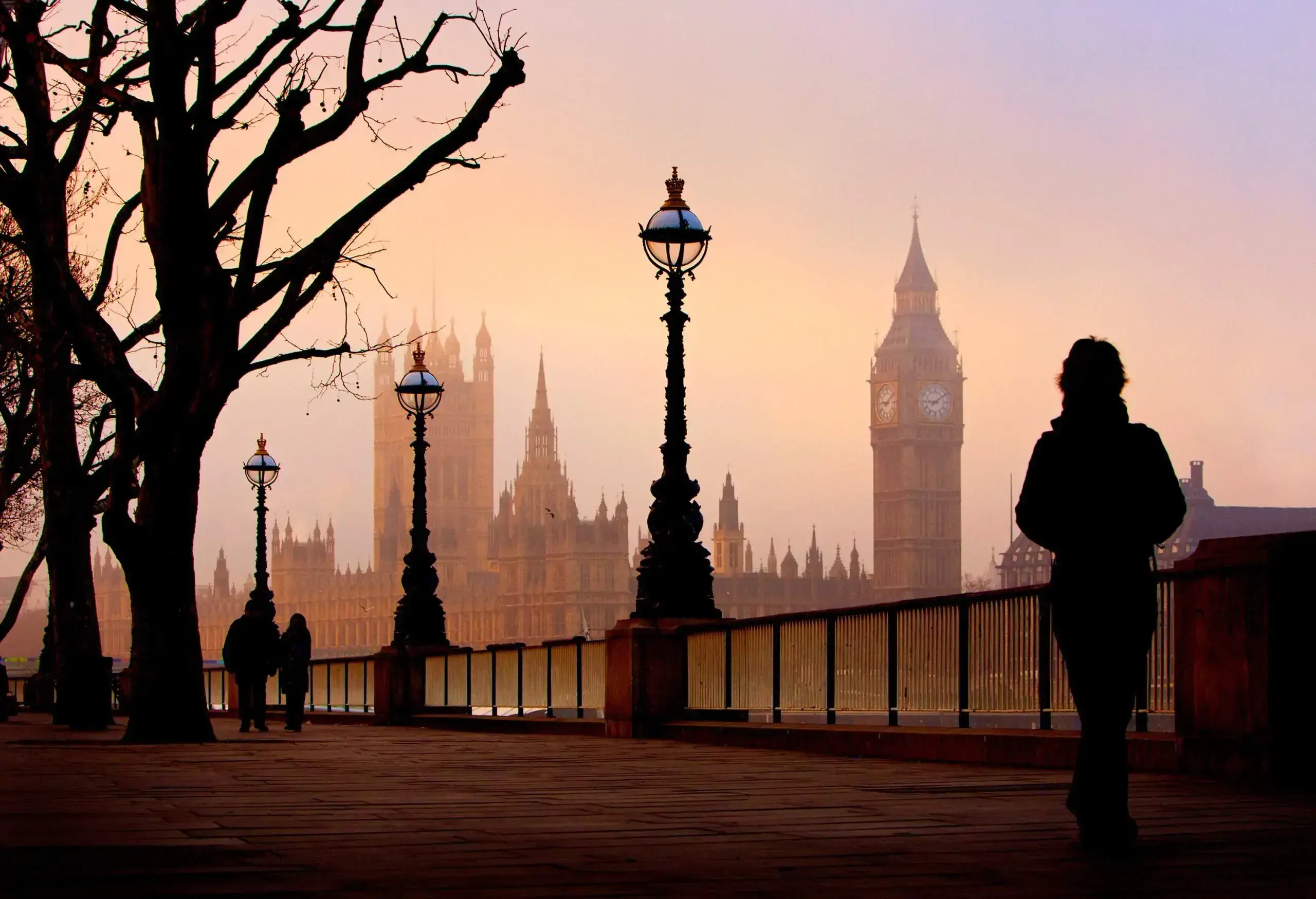 A woman's silhouette with some other tourist on a promenade lined with trees and street lights with a view of Big Ben's clock tower and the Palace of Westminster.