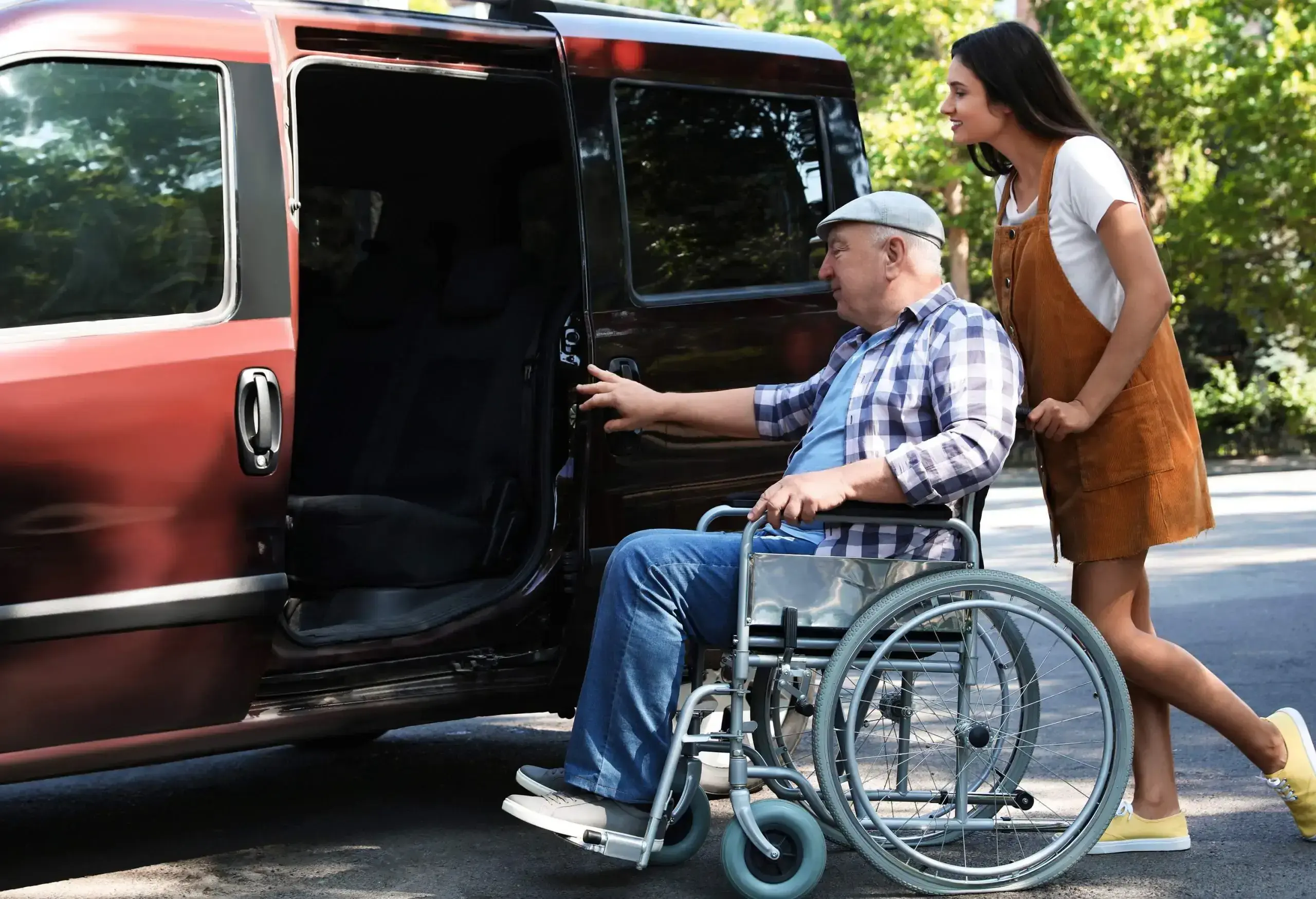 A young lady pushing a wheelchair of an elderly man to his van's door.
