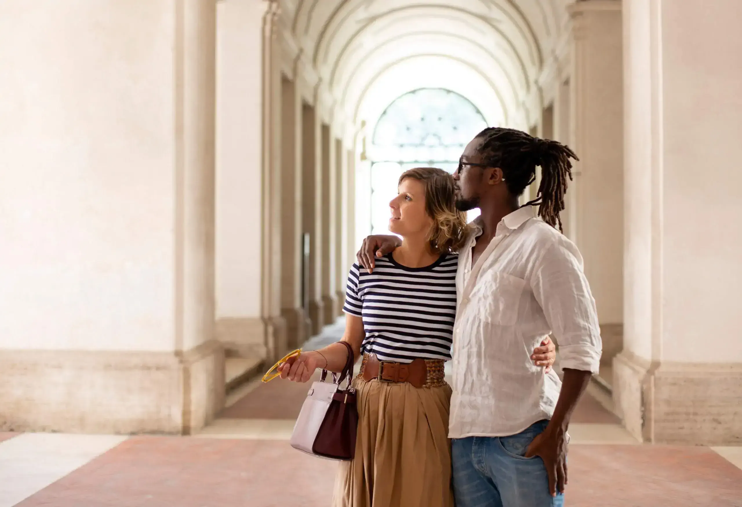 A couple stands inside a museum with archways and a beautifully decorated arch ceiling in the background.