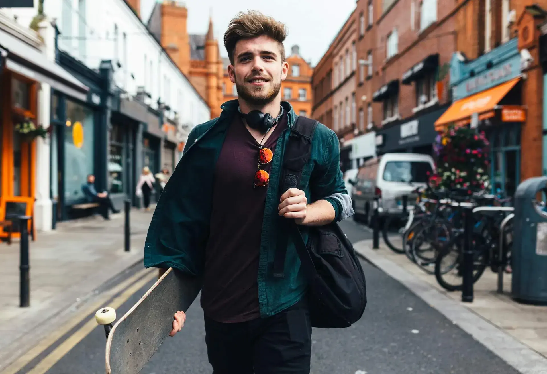 A stylish young man walking down a street, carrying a bag and a skateboard.