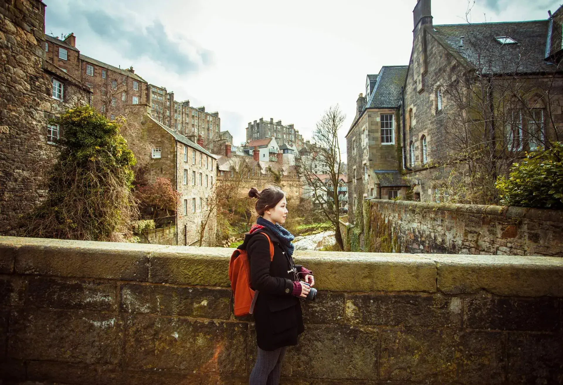 A person dressed for the cold with a backpack and a camera stands on an old bridge next to a brook surrounded by stone homes.