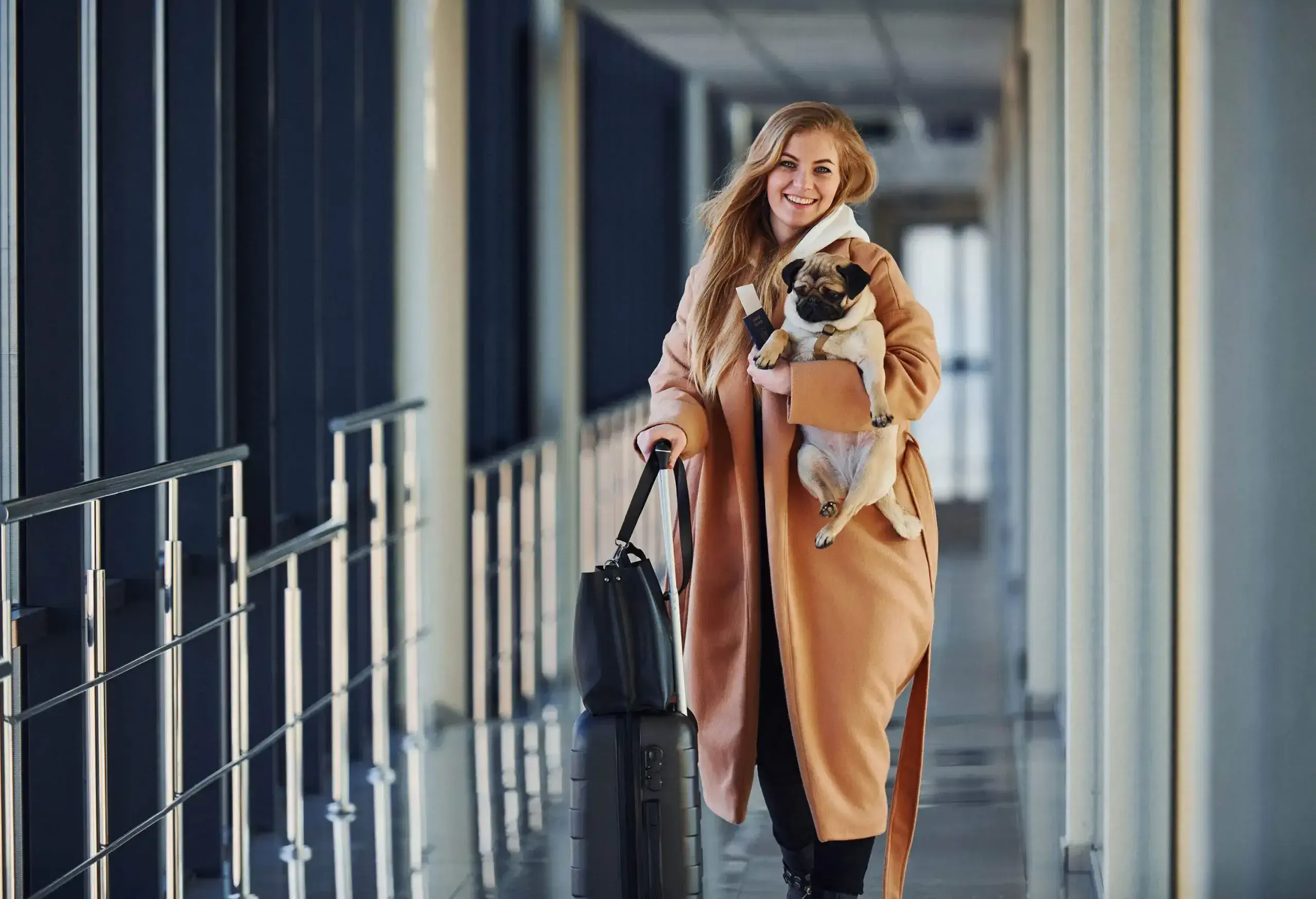 A smiling blonde woman holds her suitcase in one hand and carries her dog in the other.