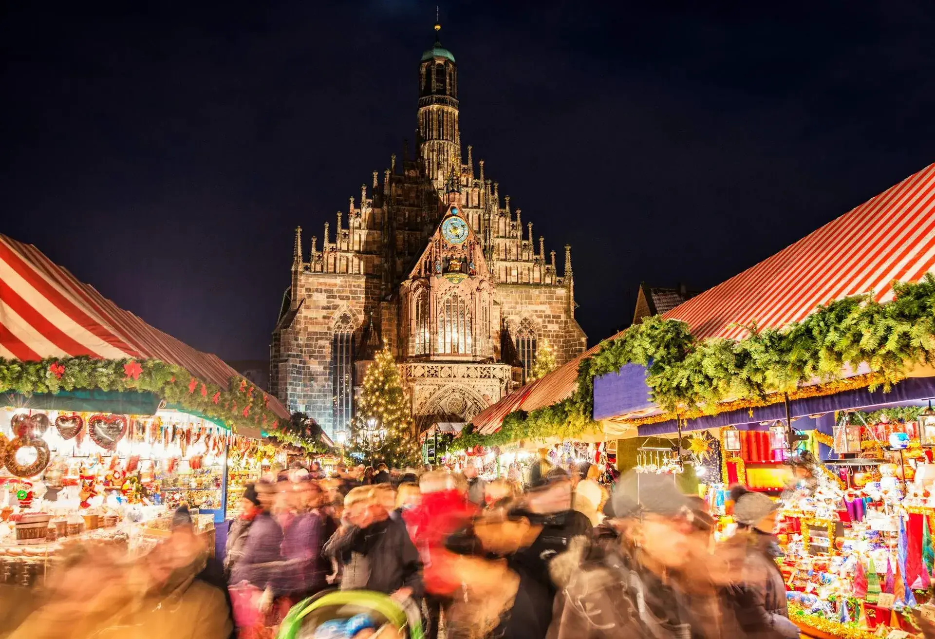 Huge crowd of people moving over Nuremberg´s world-famous christmas market (Christkindlsmarkt) at night, passing colorful illuminated christmas decoration and food stalls. Nuremberg´s landmark Frauenkirche (Church of our Lady) can be seen in the back.