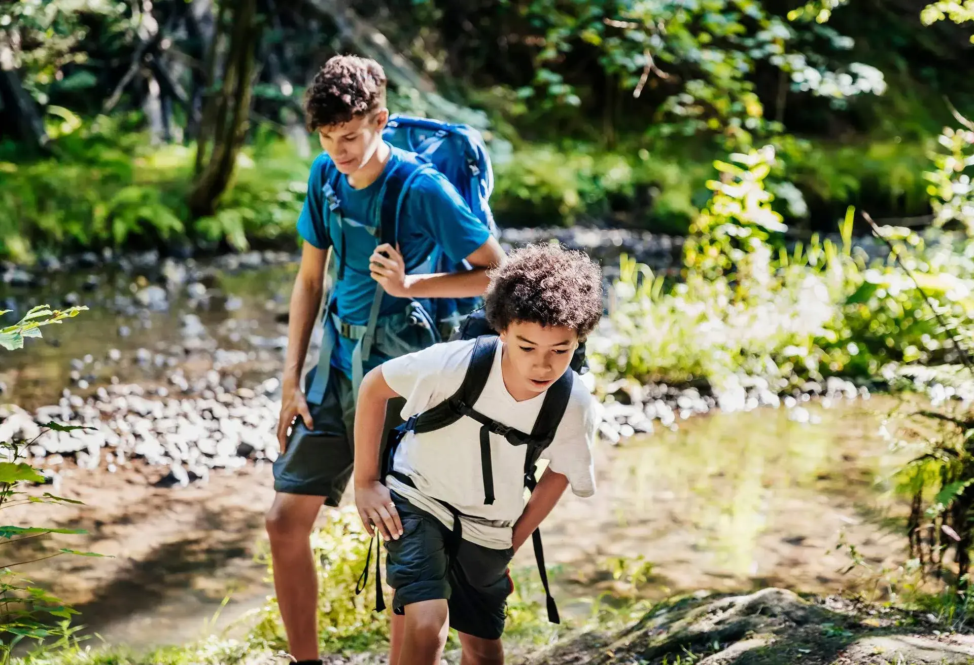 Two brothers wearing rucksacks hiking by a river together while navigating a woodland trail.