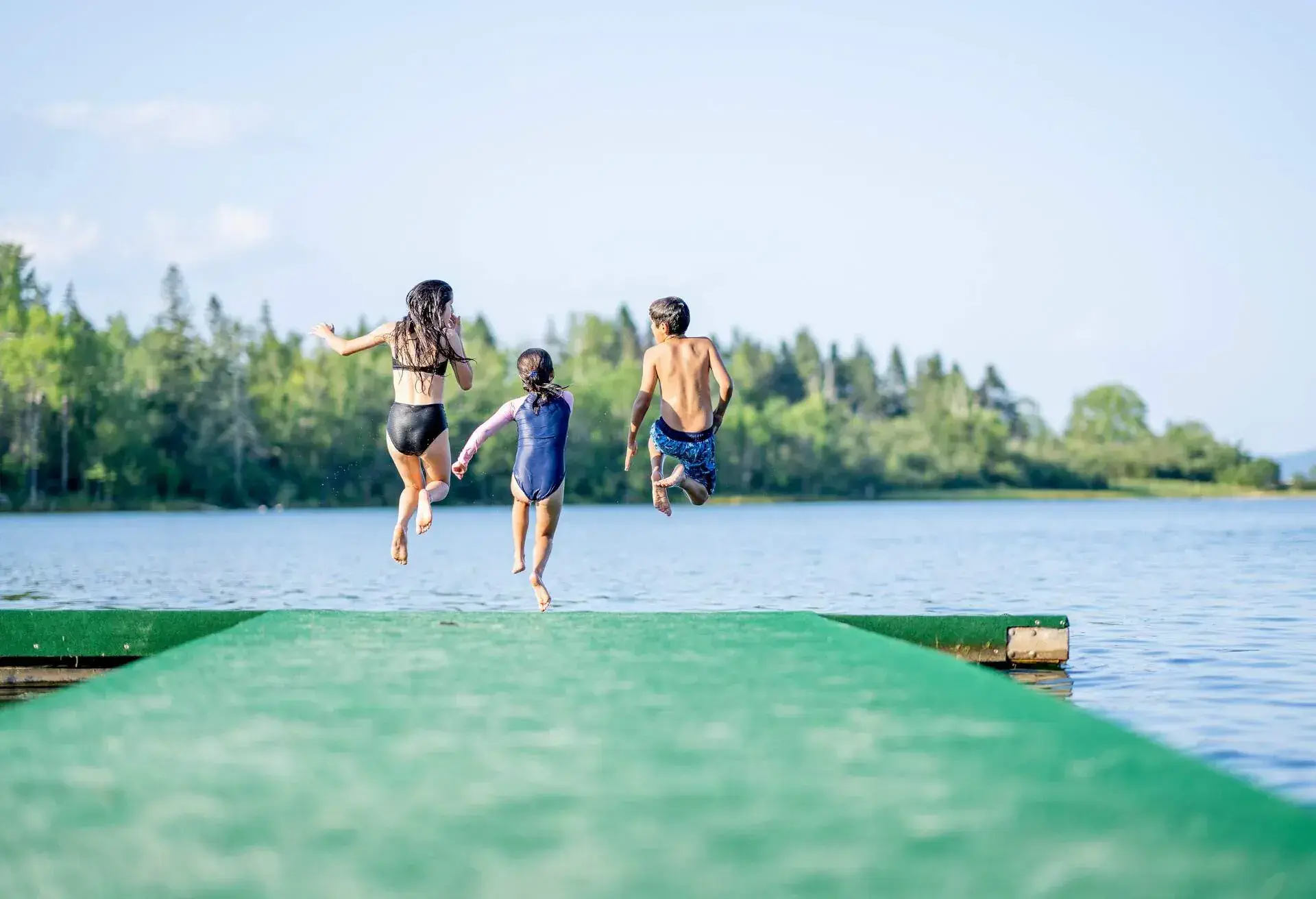 A small group of three children are seen in mid air jumping off the end of a dock on a sunny summer day. They are each striking a fun pose before they hit the waters surface to make a splash.