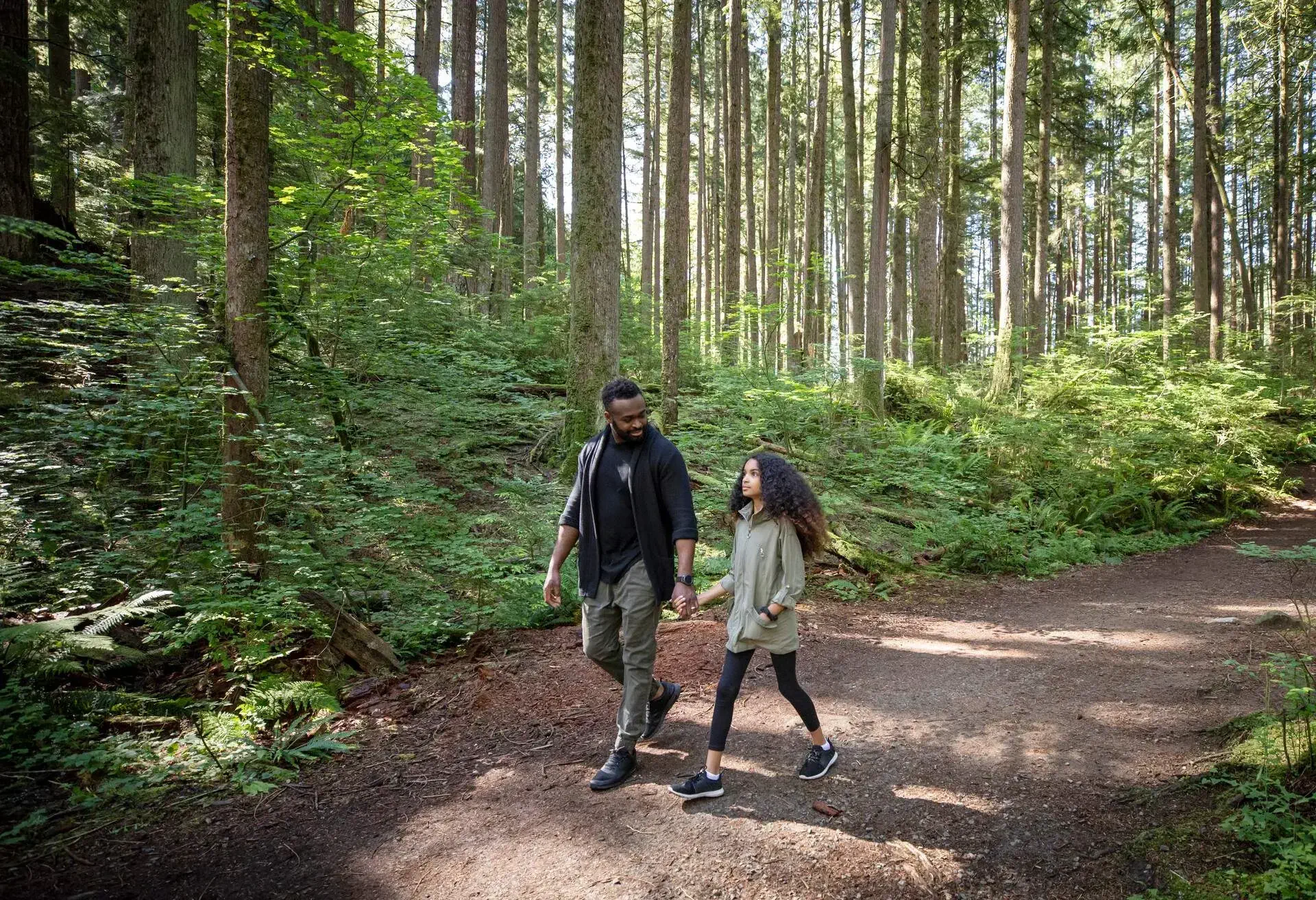 A man holds his curly-haired daughter's hand as they walk together on a sunlit forest trail
