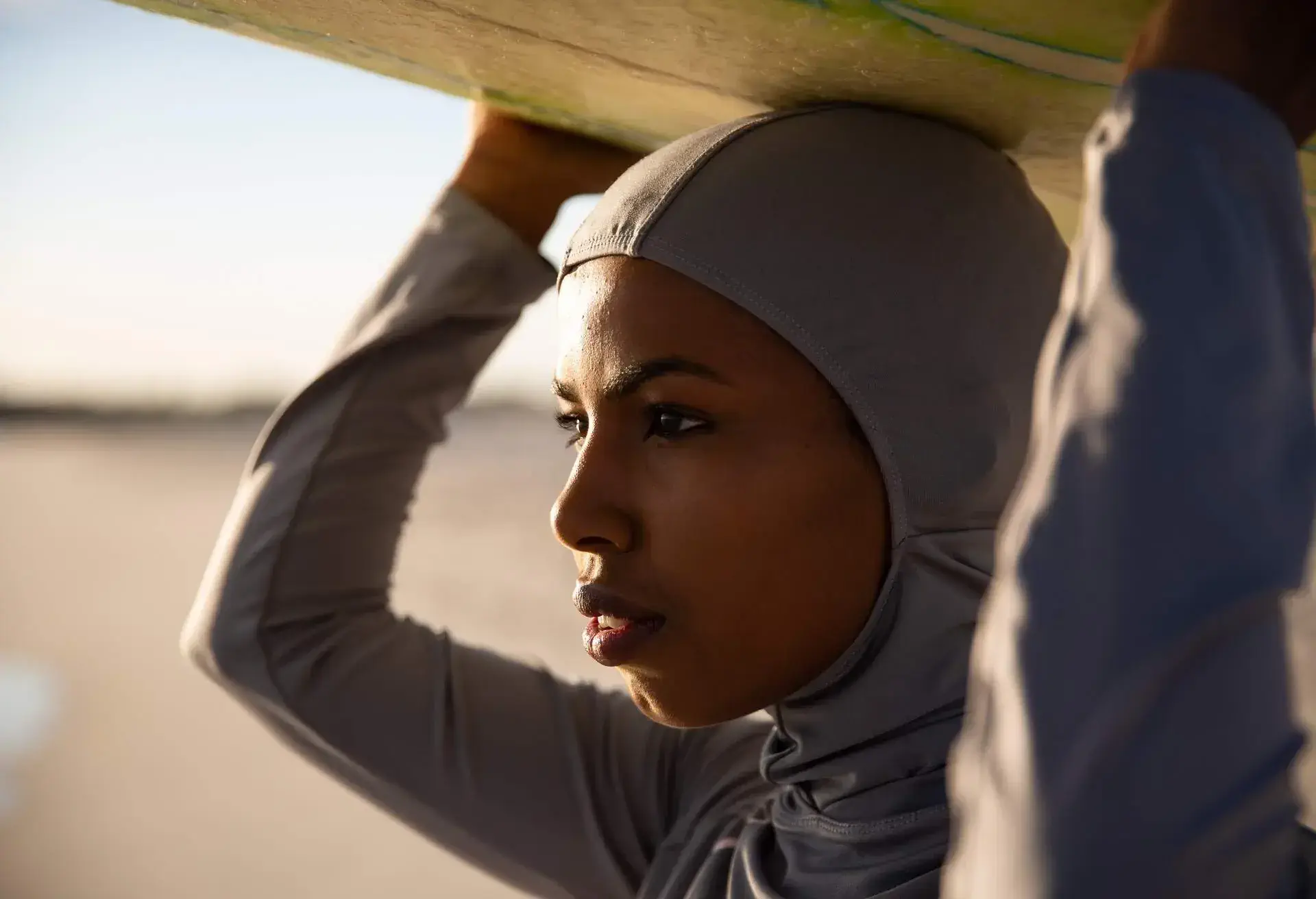 Australian muslim girl of African heritage wearing hijab and looking at distance while holding a surfboard