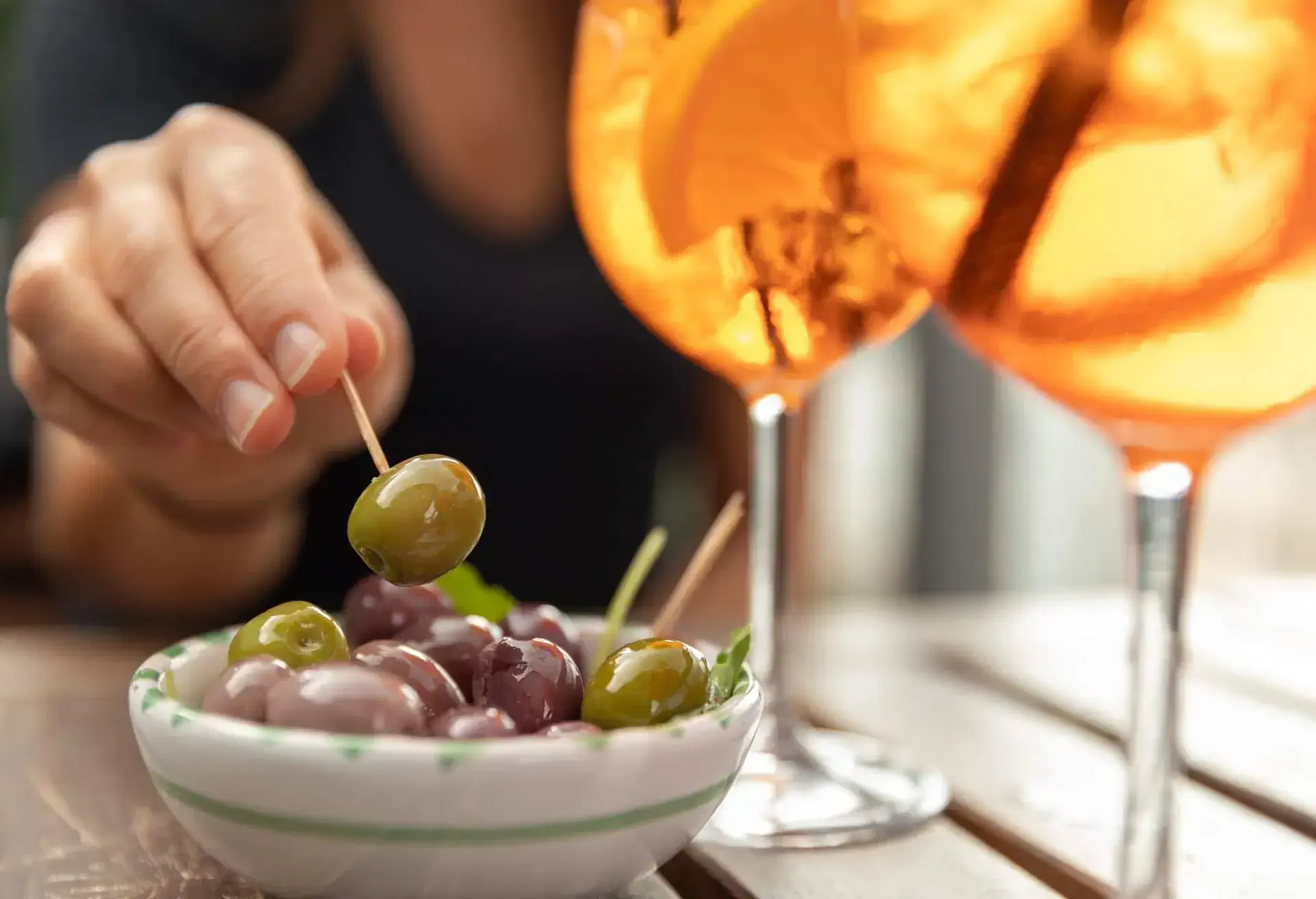 A bowl of olives and two drinks are standing on a table on a rooftop bar in Copenhagen. A woman is sitting by the table and she's picking up an olive with a toothpick.