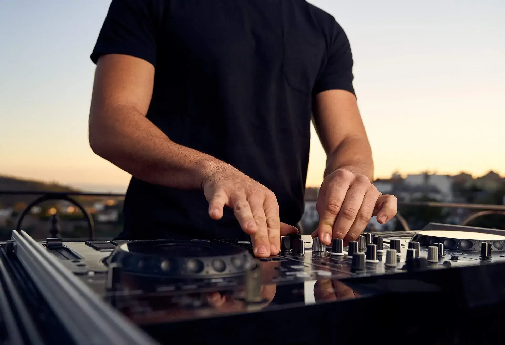 Unrecognizable male DJ hands playing music in a session with a mixer at a sunset party on a terrace. Electronic music concept.
