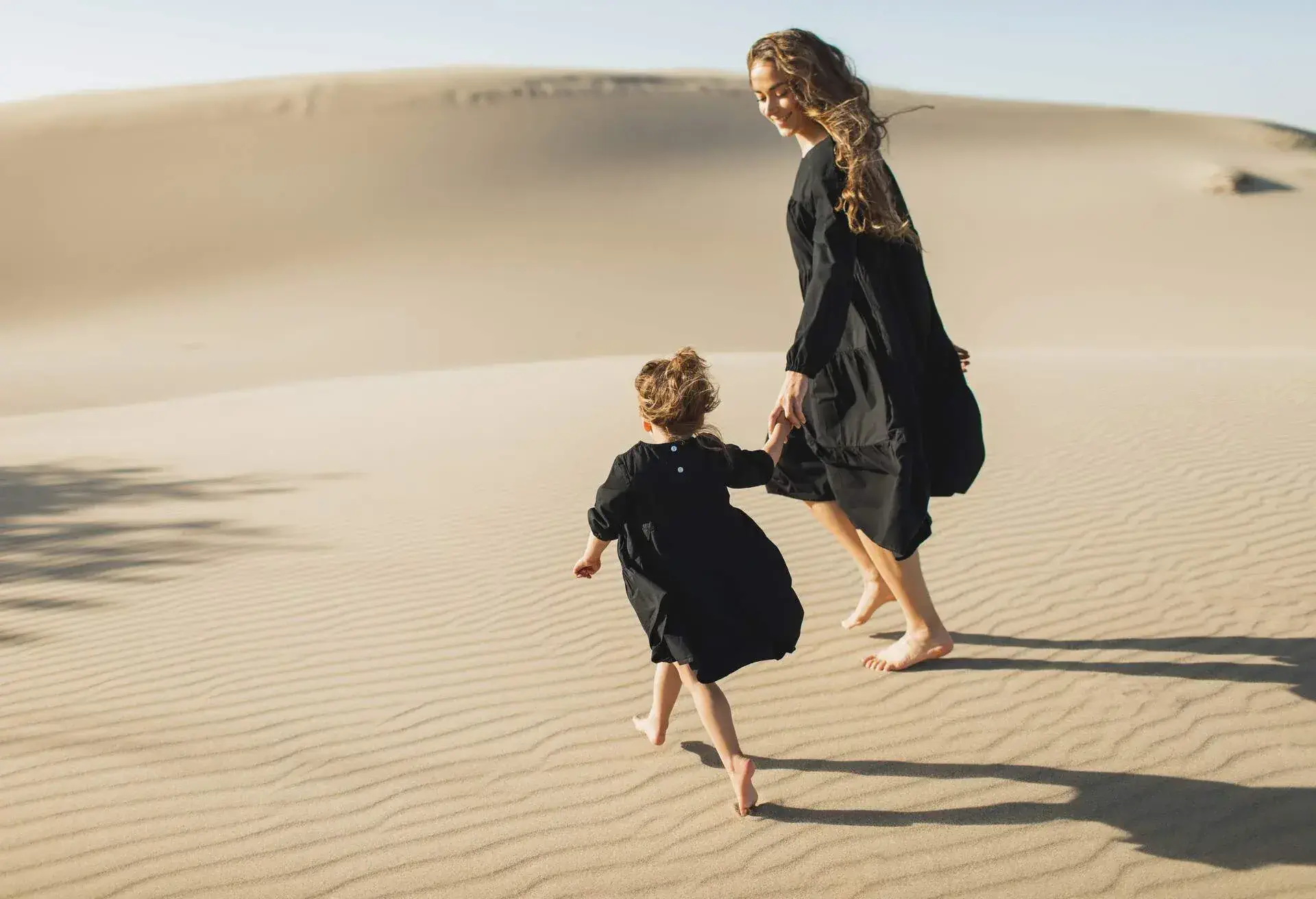 A mother and daughter strolling barefooted on sand dunes in the desert.