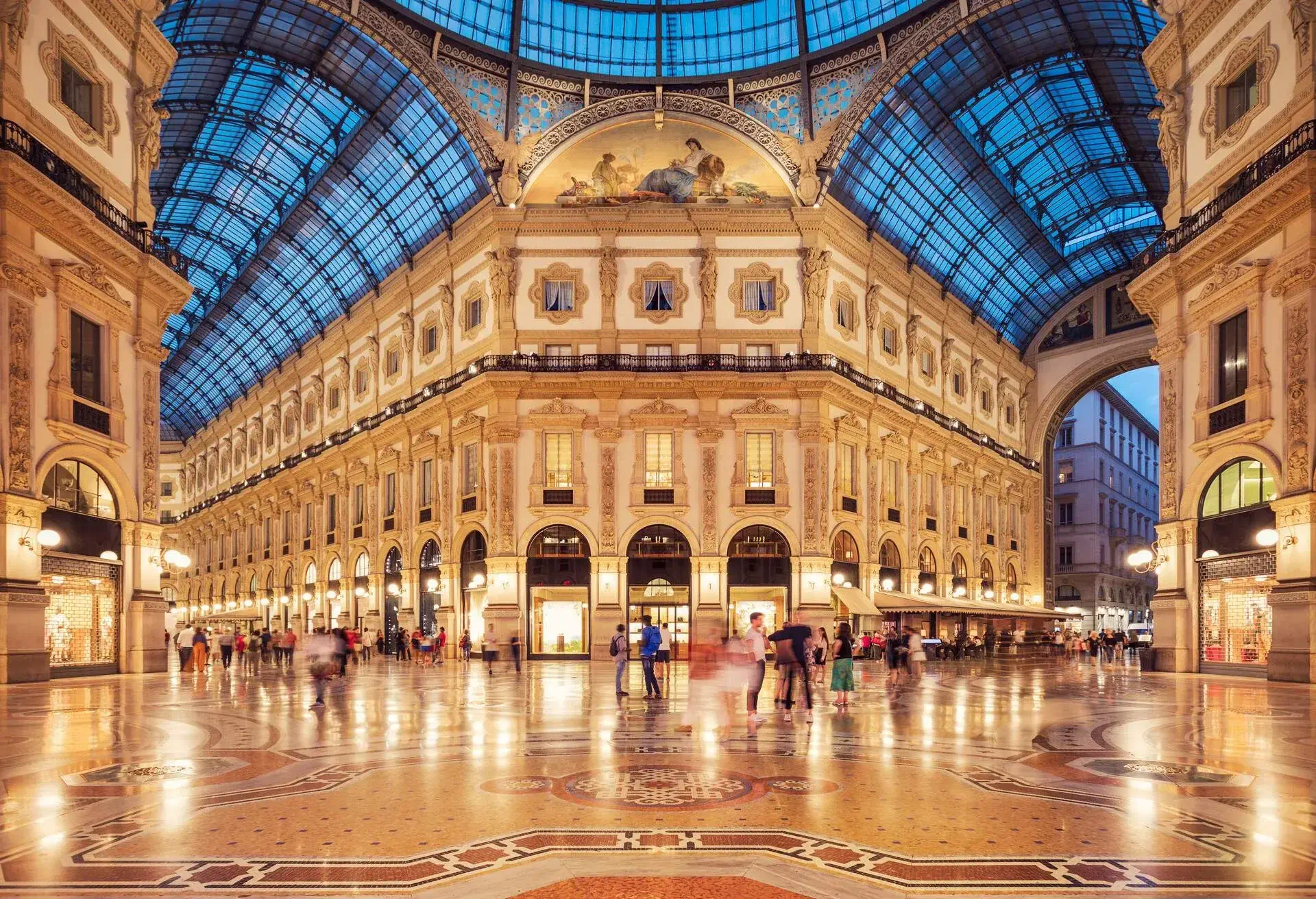 An elegant and historic indoor shopping gallery with blue glass ceilings, arched shop doors, paintings and windows with intricate carvings.