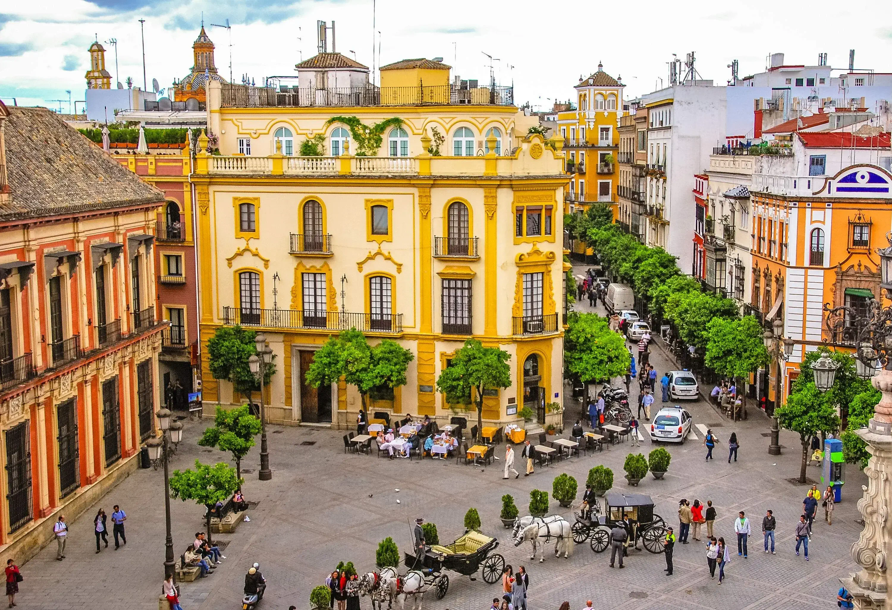 A crowded plaza with horses strapped on carriages surrounded by outdoor seating areas along the colourful city buildings.