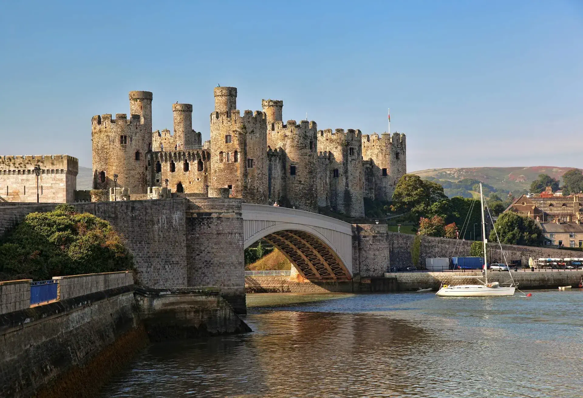 The front of a castle atop a rocky coastal ridge behind a bridge over the river.