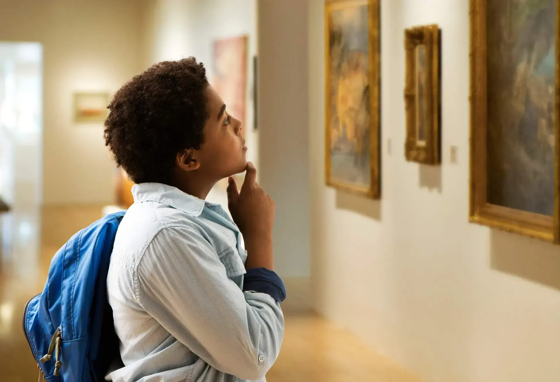 A person with a backpack gazes at a painting, hands thoughtfully resting on their chin, as they appear to be contemplating the artwork's meaning.